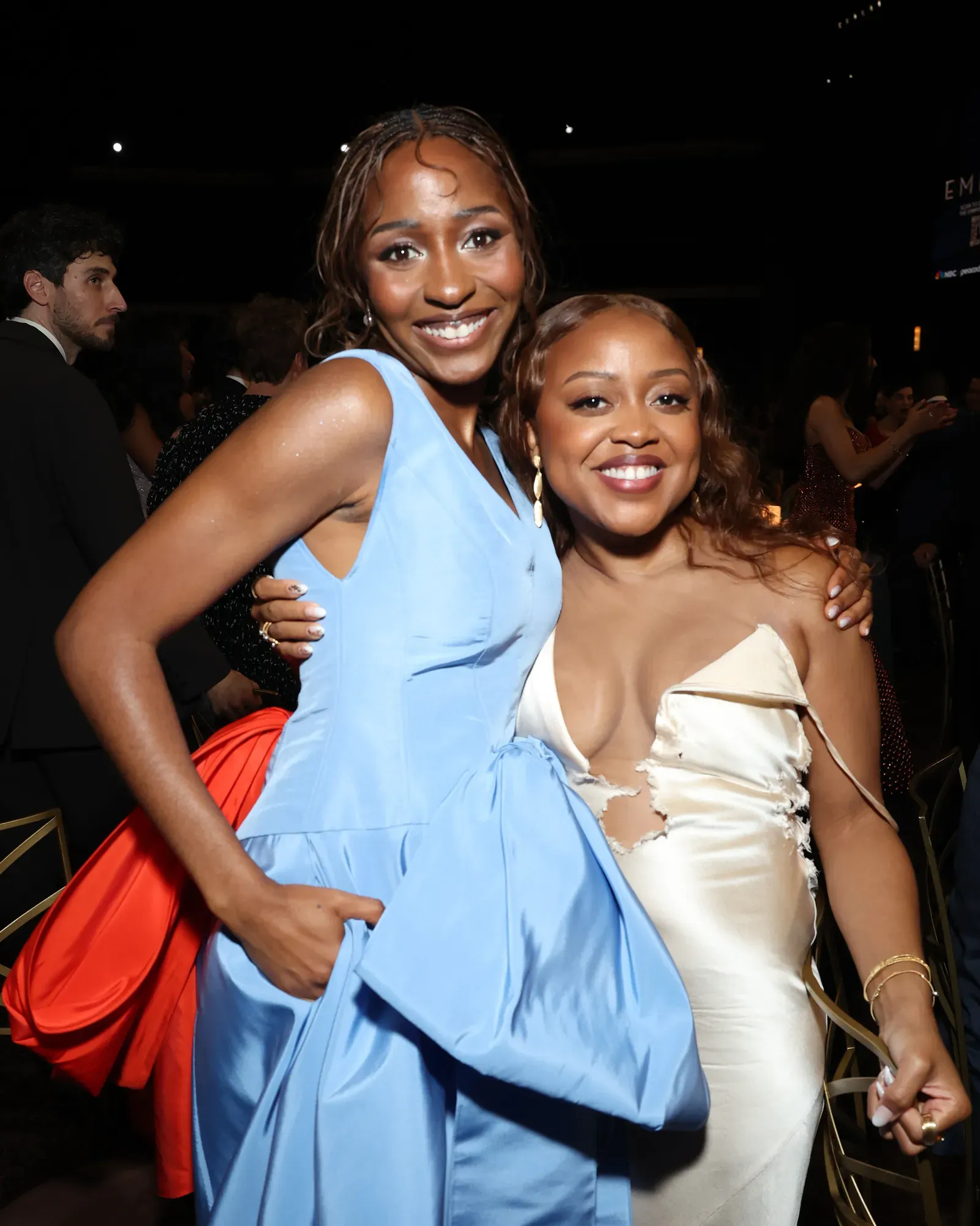 Two women pose together at a formal event, smiling for the camera; one in a light blue dress, the other in an ivory dress with ripped details, against a dark background.