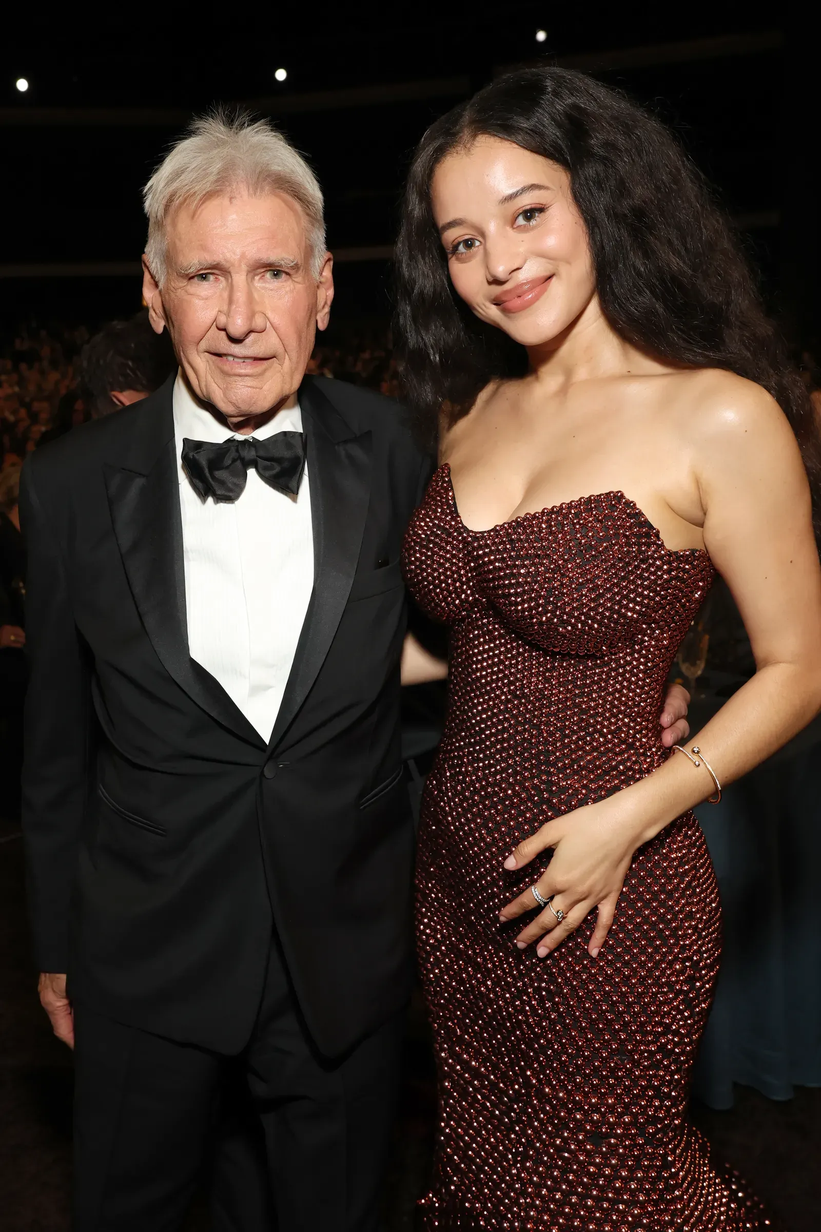 Older man in a black tuxedo with a bow tie standing next to a smiling woman in a sparkling red gown at a formal event.