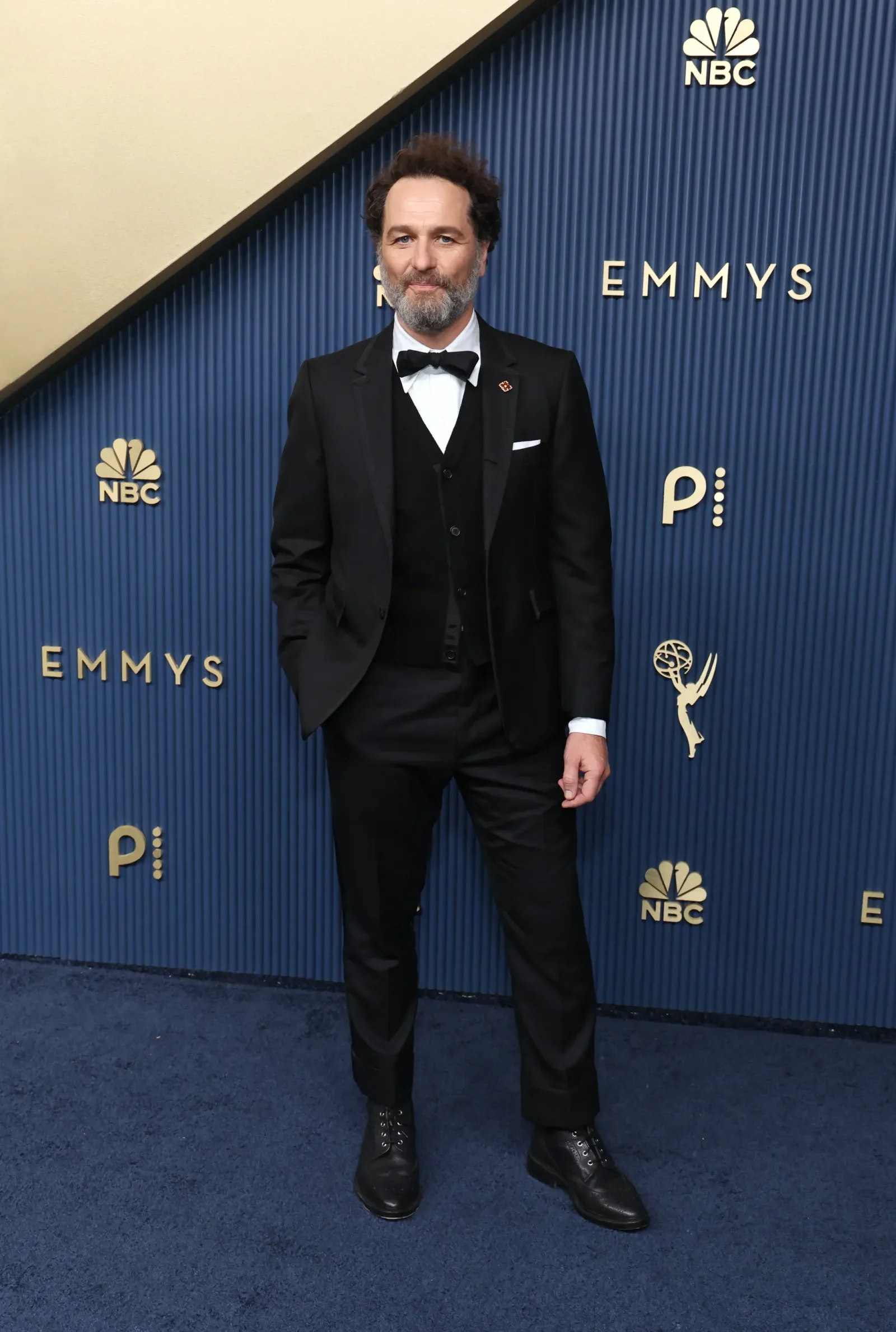Man in a black tuxedo with a bow tie posing on a blue Emmy Awards backdrop with NBC logos behind him.