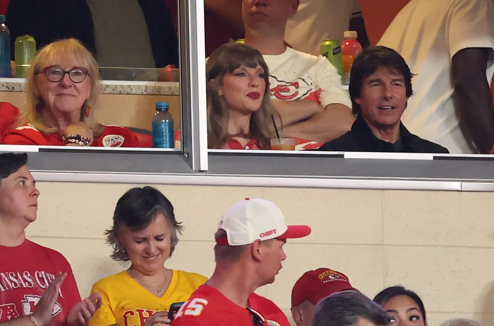 Three spectators in a stadium box watching a game: a blonde woman with glasses on the left, a gray-haired woman with red lipstick in the middle, and a dark-haired man on the right; foreground fans wear Chiefs gear.