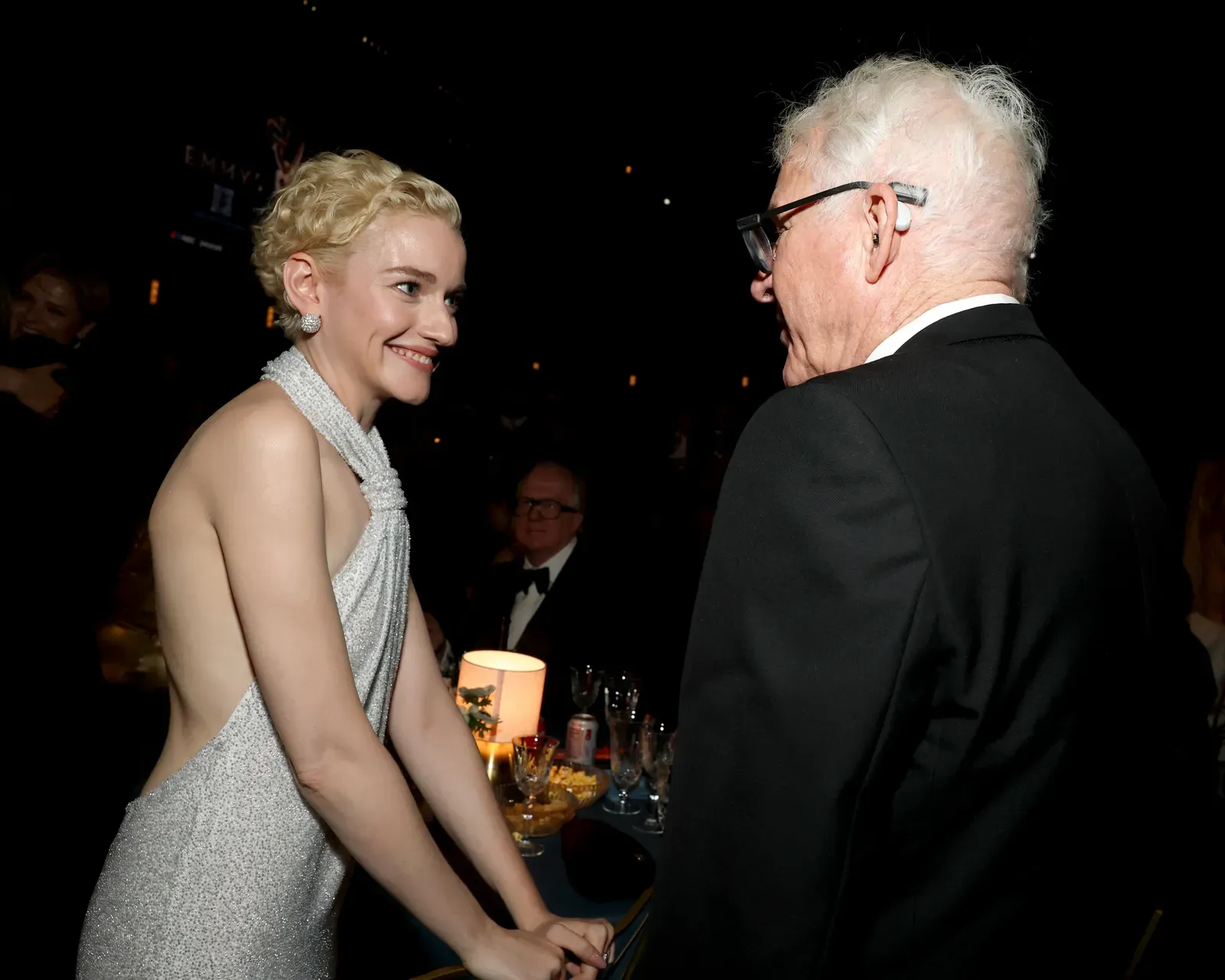Smiling woman in a silver glitter gown shakes hands with an older man in a black suit at a formal event, candlelit tables in the background.