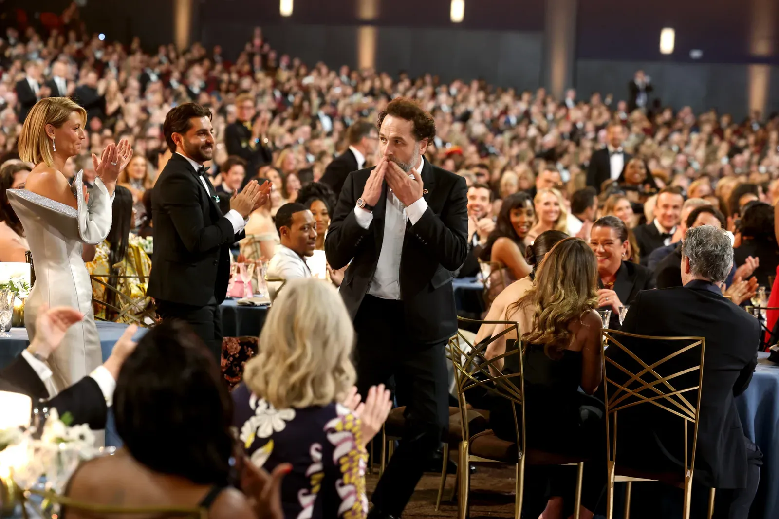 Crowd at a formal awards dinner applauds as a man in a suit walks through the tables, covering his mouth in surprise or emotion.