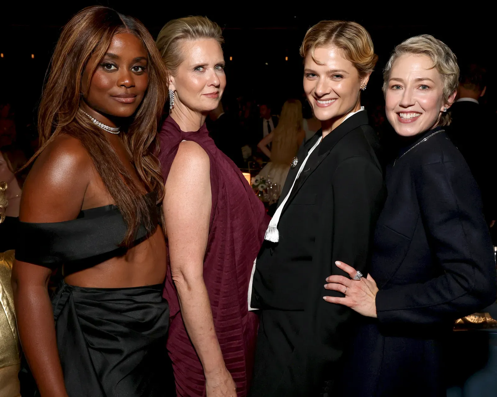 Four women in formal attire posing together at a dim event, smiling at the camera.