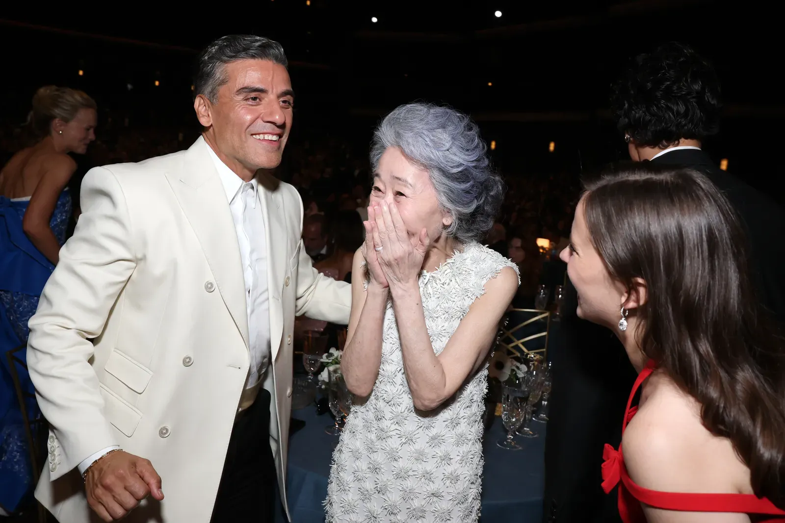A man in a white suit chats with an elderly woman in a silver floral dress, both smiling at a formal event.