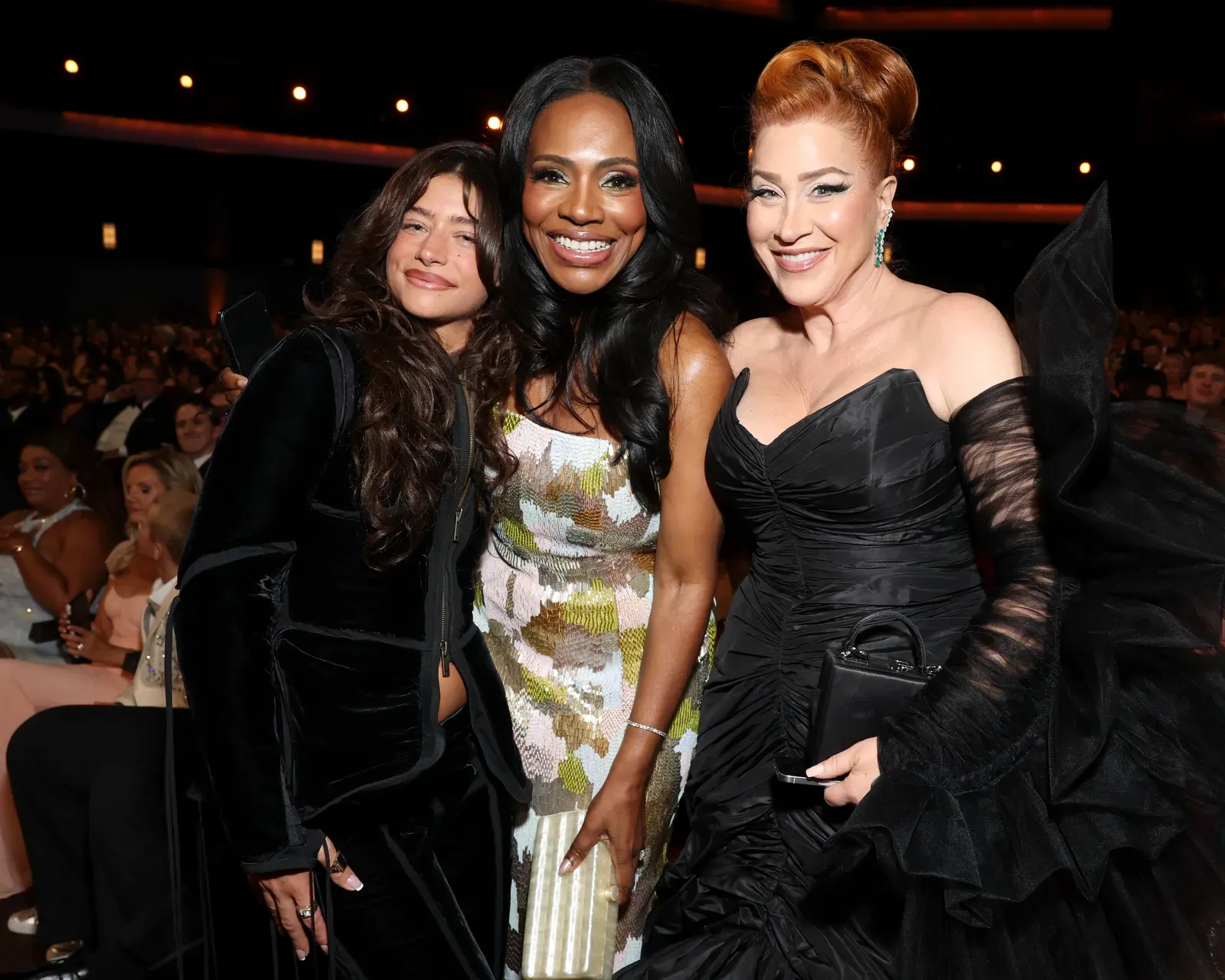 Three women pose together at a formal event, smiling for the camera with a seated audience in the background.