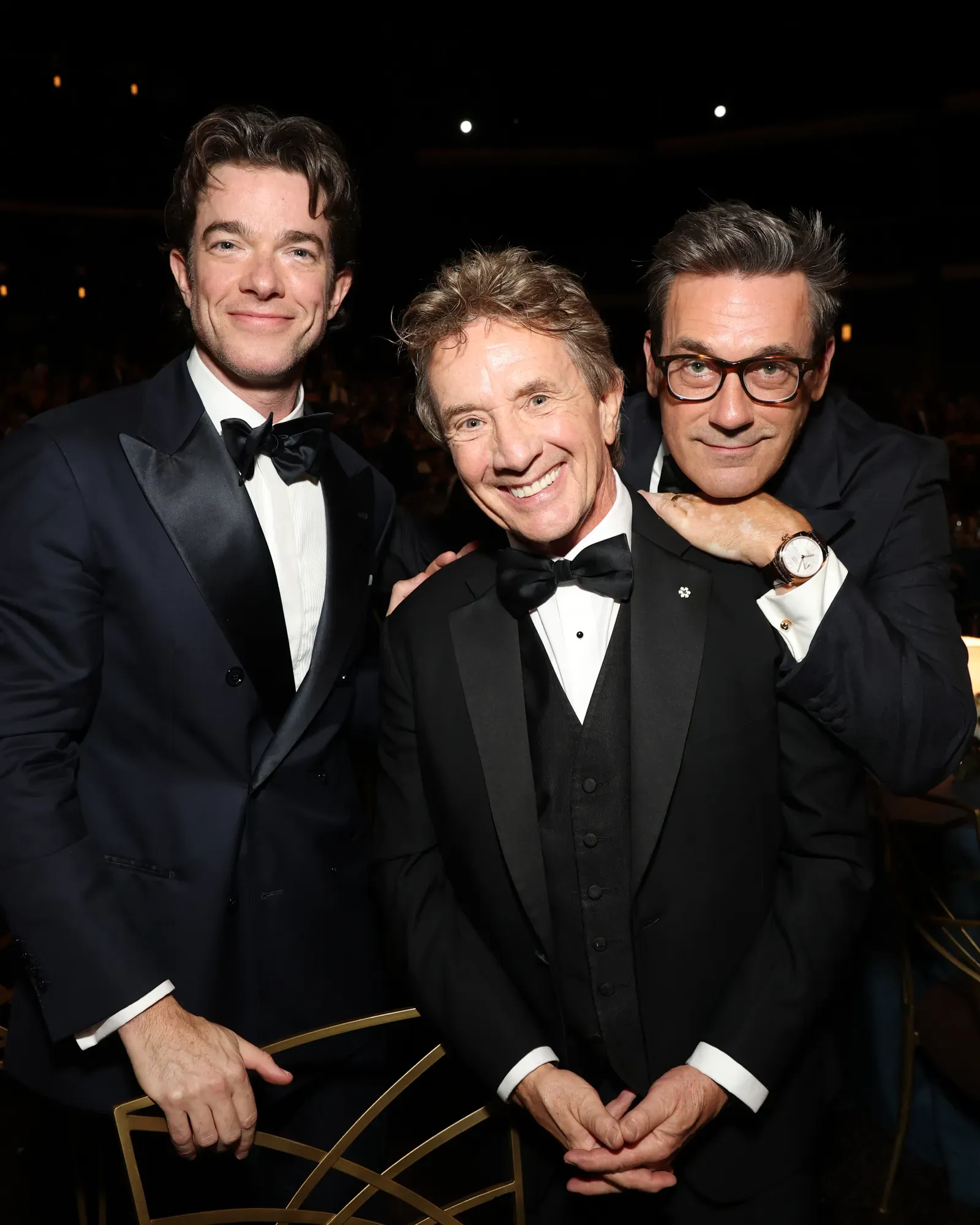 Three men in tuxedos posing together at a gala, smiling for the camera beside a gold geometric sculpture.