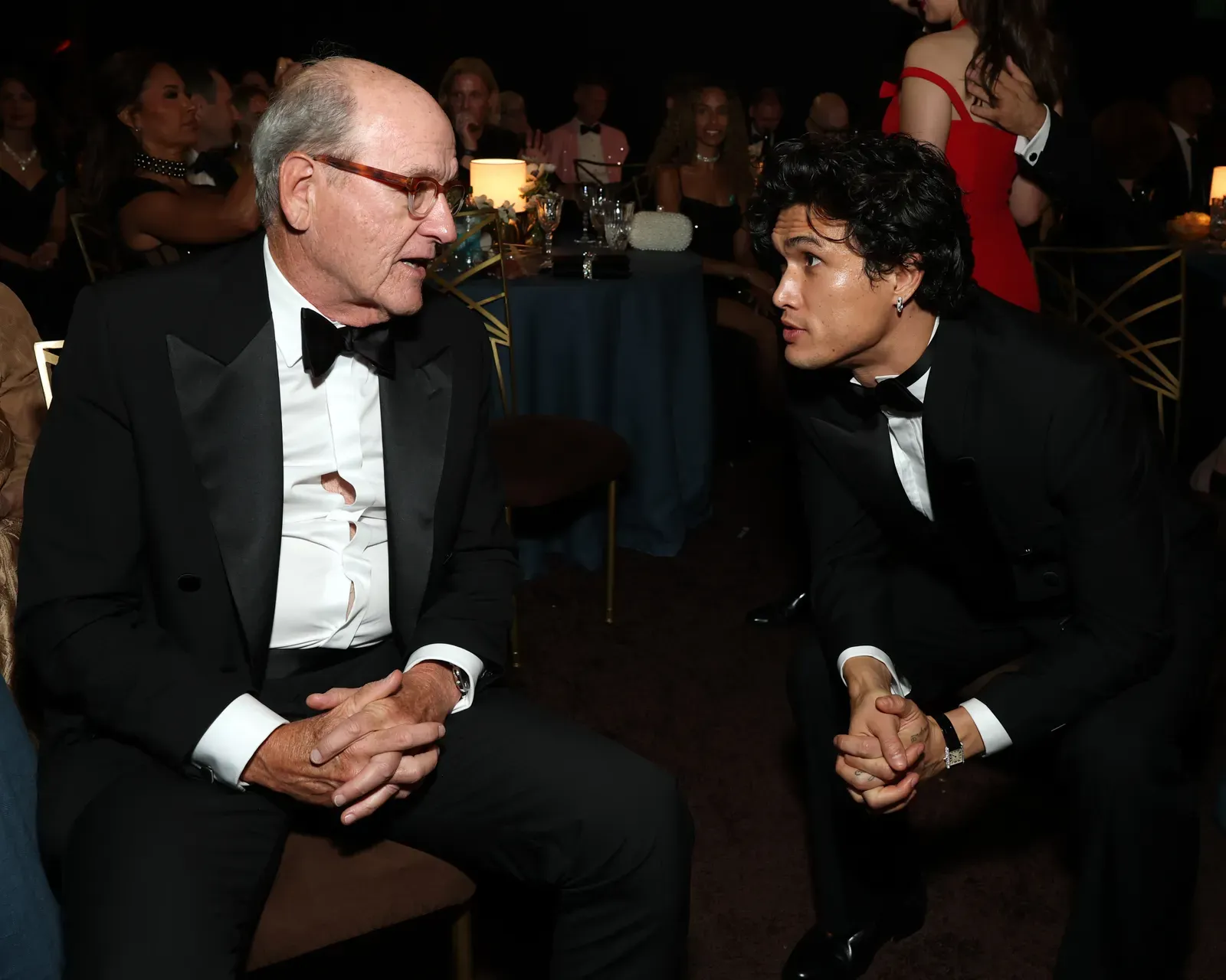 Two men in black tuxedos converse at a formal dinner, one older with glasses on the left and a younger man leaning in on the right, with a candlelit table in the background.