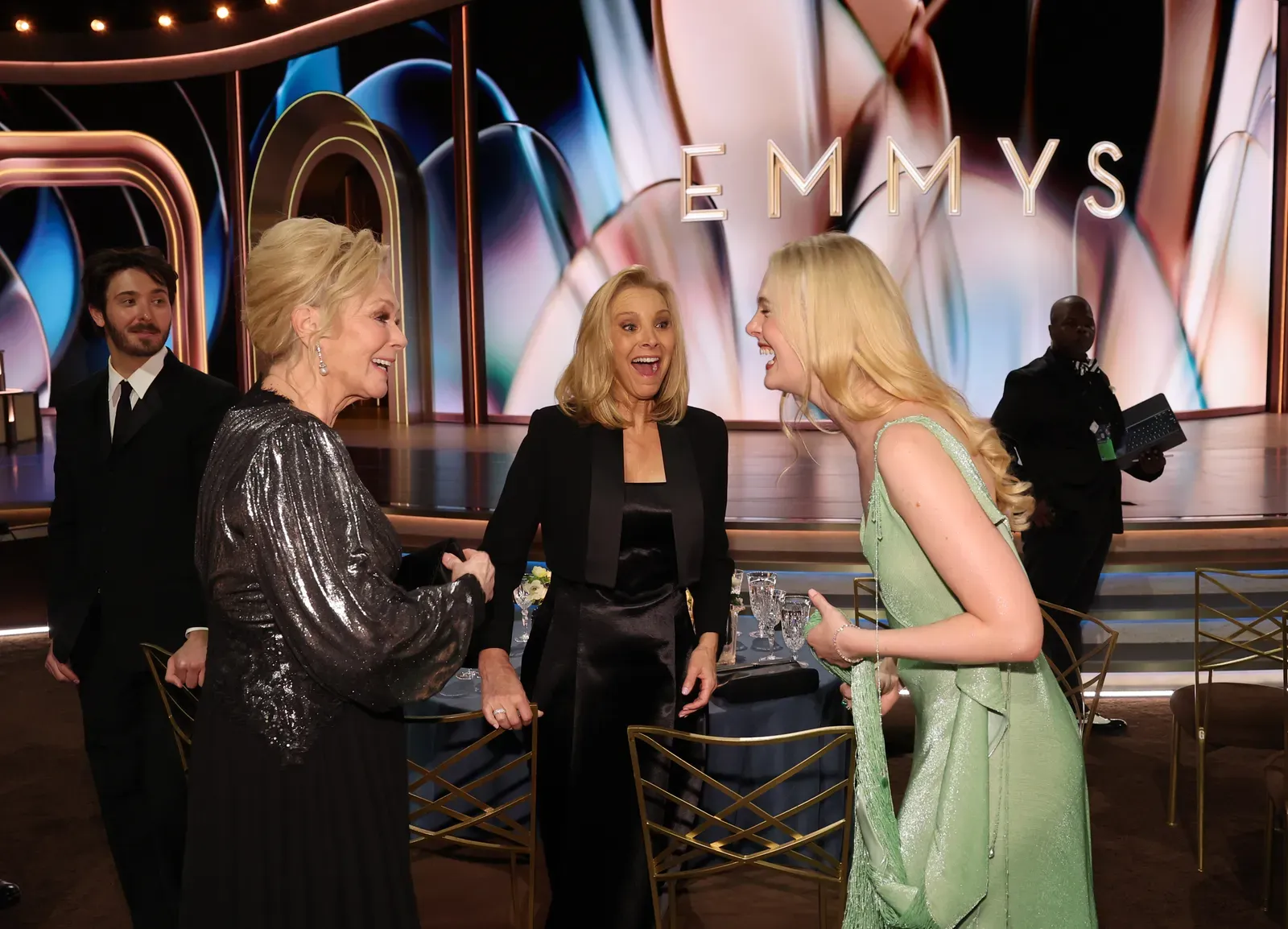 Three women in elegant gowns greet each other at an Emmy Awards event, with a colorful stage backdrop that reads EMMYS behind them.