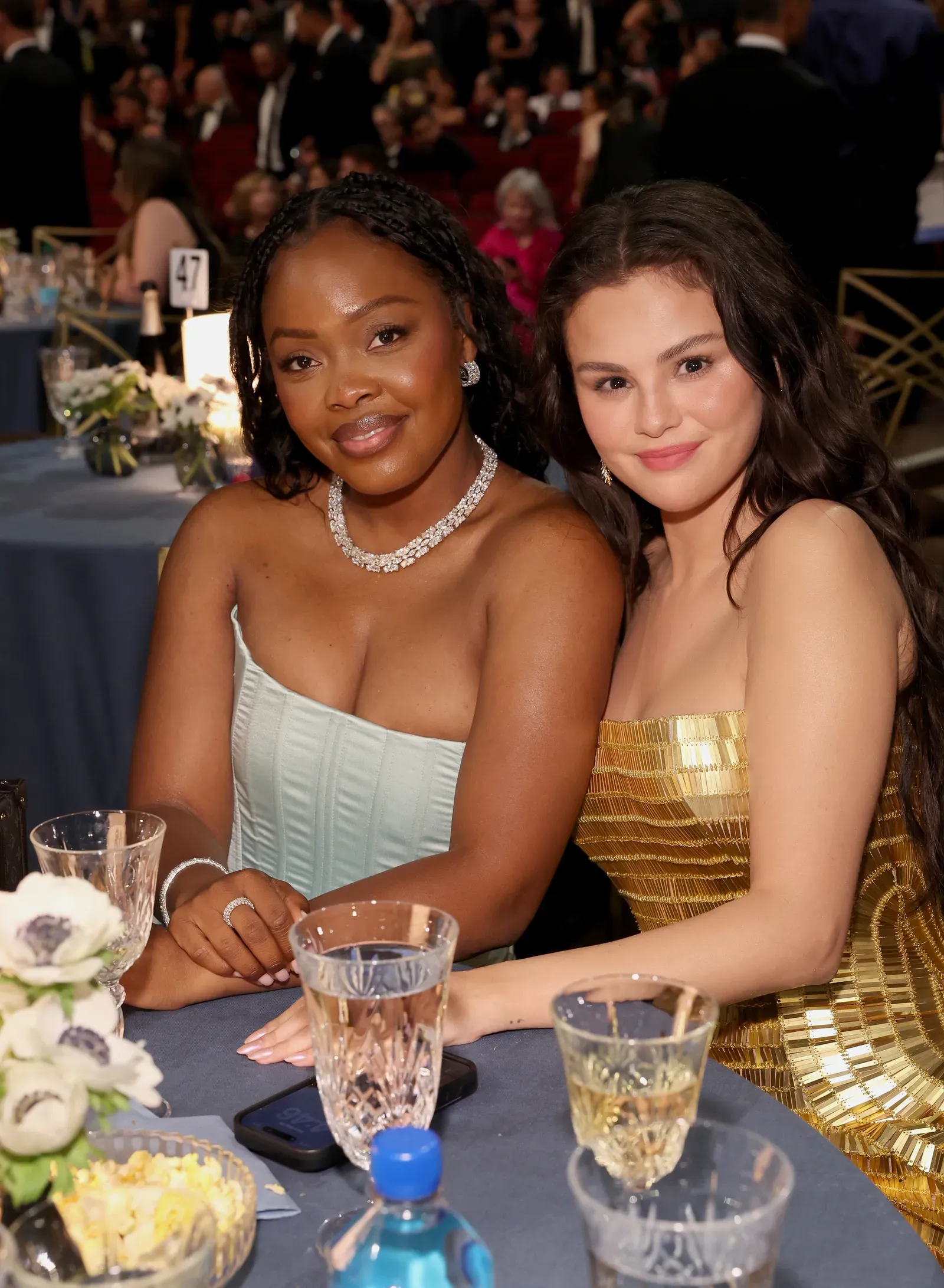 Two women in formal gowns sit at a banquet table, smiling at the camera; glasses and flowers on the table in front of them.