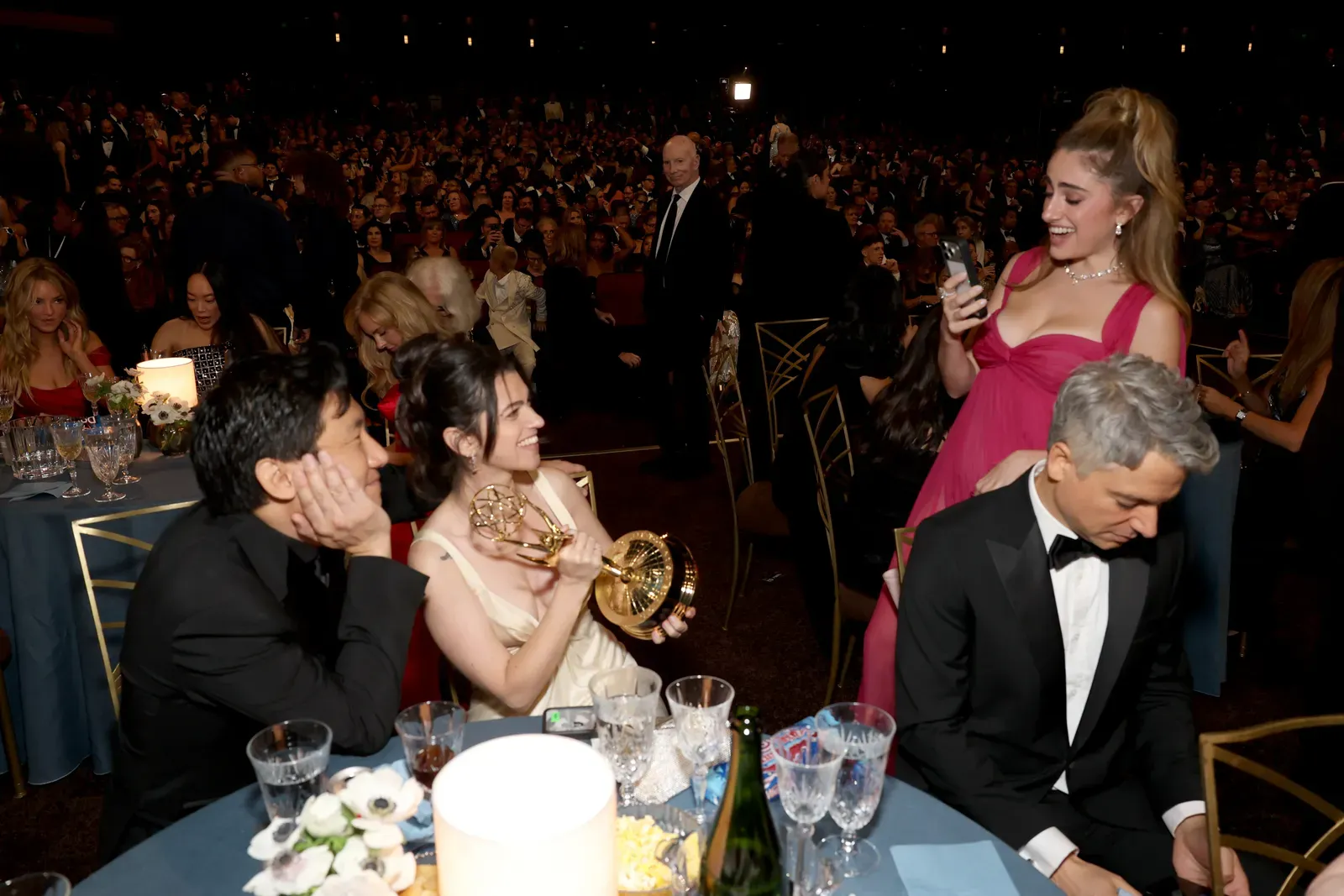 Guests seated at a formal awards banquet; a woman in a pink dress stands and smiles at her phone while others chat and laugh at the table.