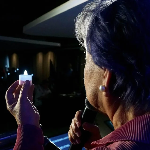 Person with short purple hair, seen from behind, holds a lit lighter near a candle in a dark room.