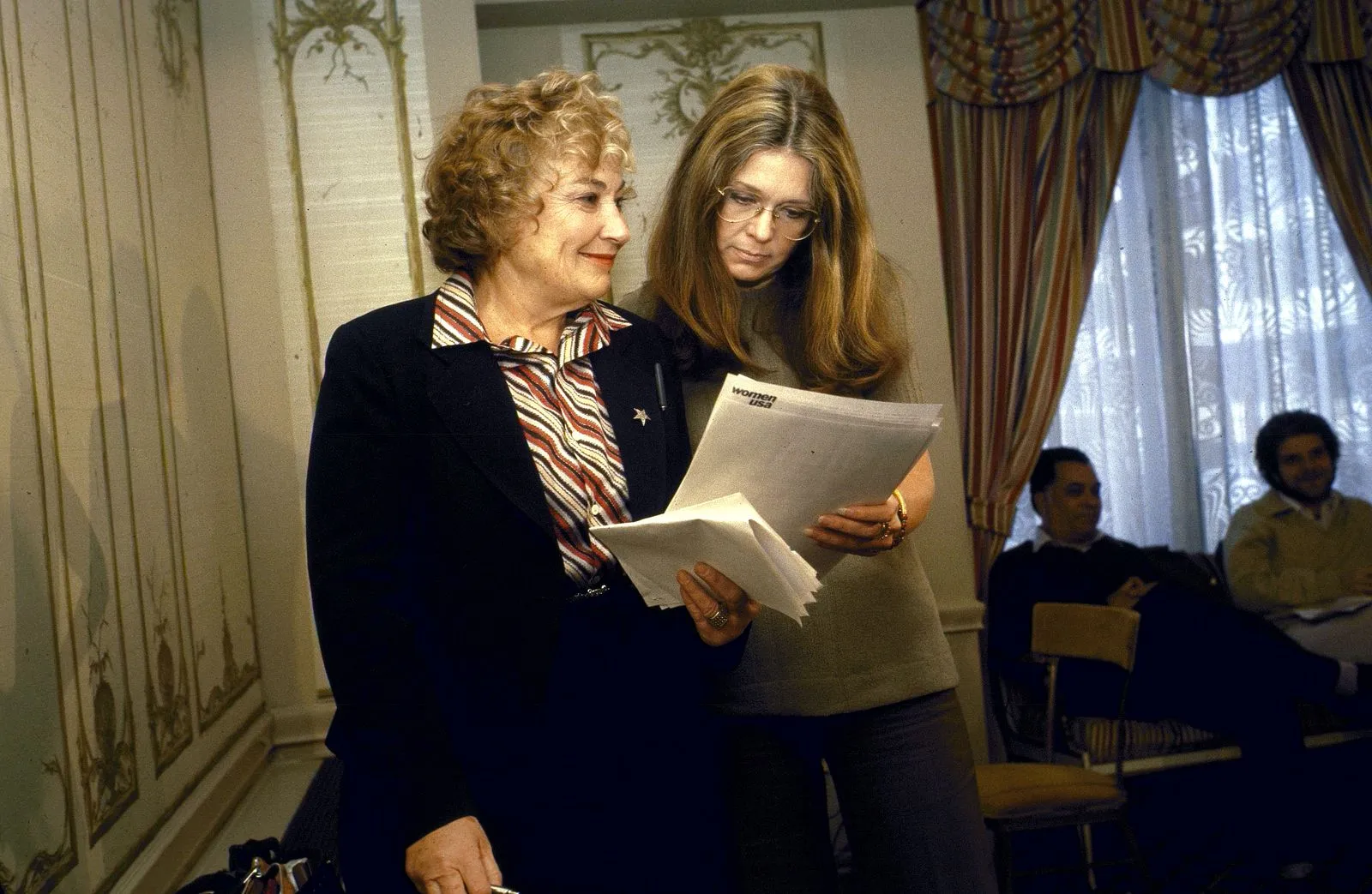 Two women stand and review documents together in a formal room, with others seated in the background.
