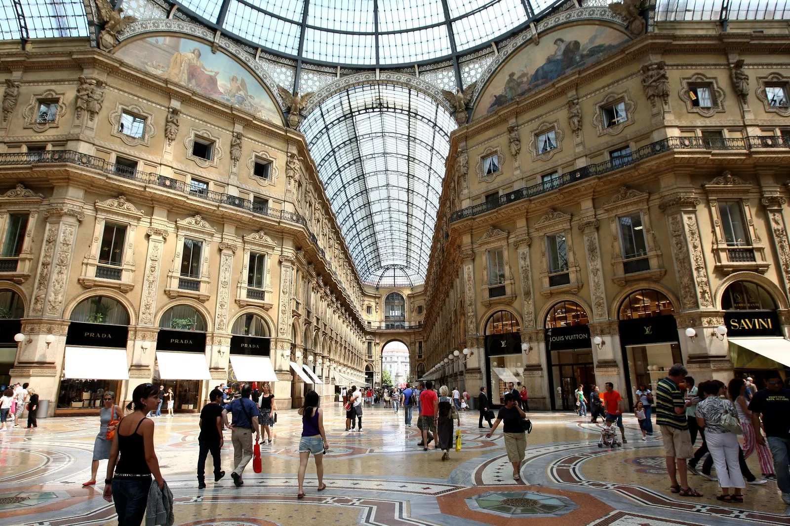 Interior of a grand 19th-century shopping arcade with ornate stone arches, glass roof, and luxury shops; shoppers walk on a mosaic floor.