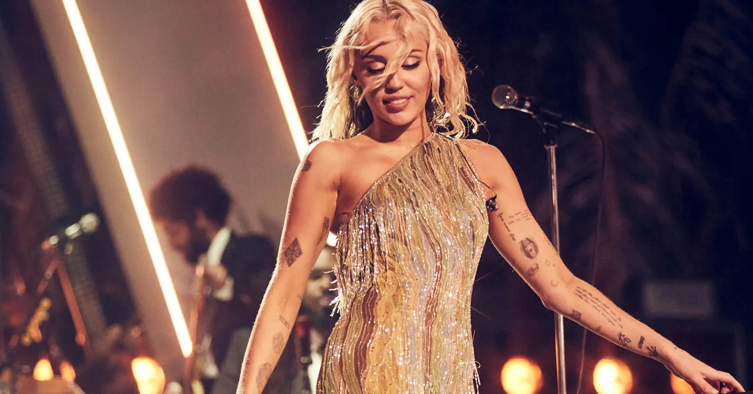 Female singer in a glittering gold dress performs on stage with arms outstretched, tattoos visible on her arms.