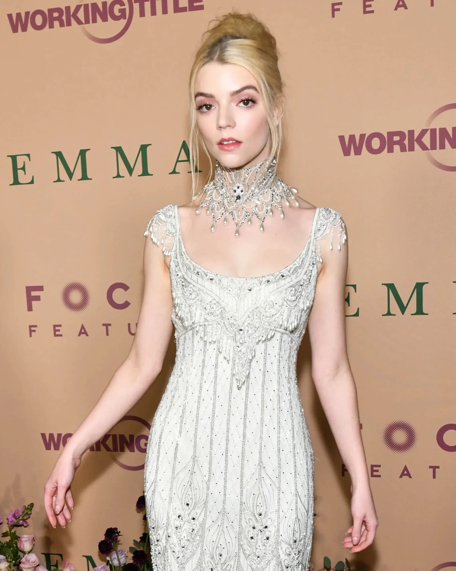 Blonde woman in an intricate silver beaded gown with a jeweled high necklace posing on a peach backdrop at a fashion event.