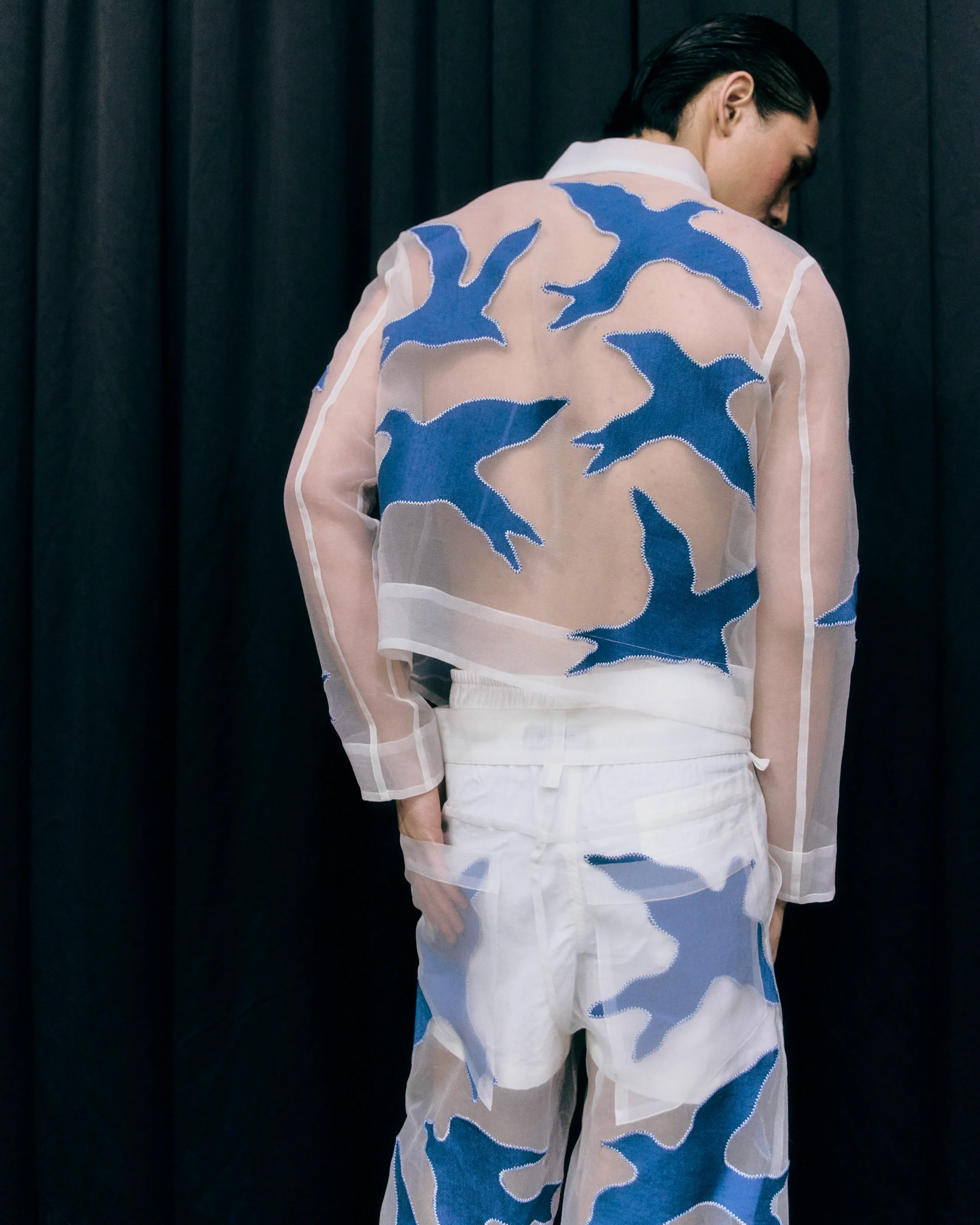 Back view of a person wearing a sheer pink shirt with blue camouflage patches against a dark curtain backdrop.