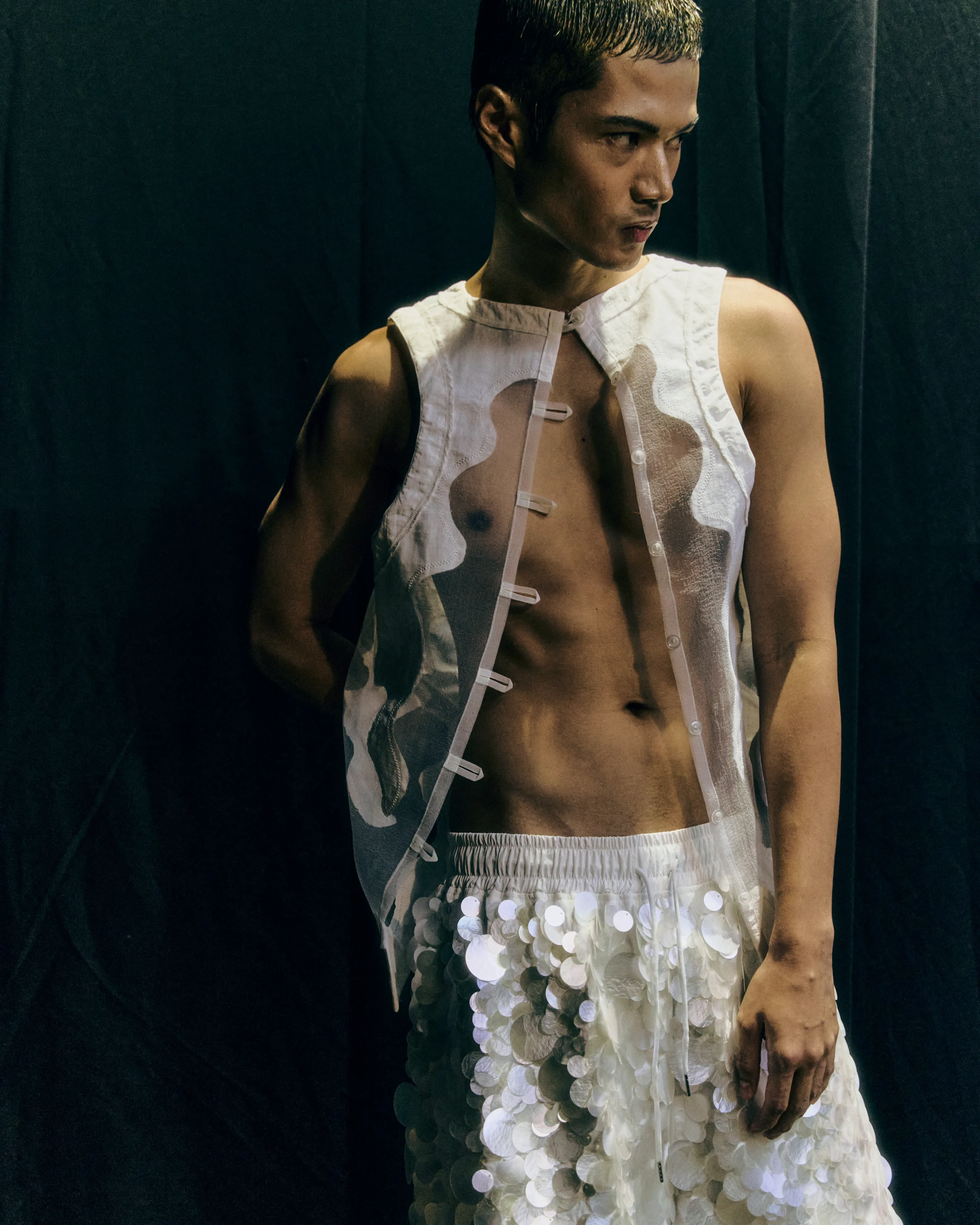 Man wearing a white sleeveless vest with decorative closures and a sequined skirt, standing against a dark background, looking to his left.