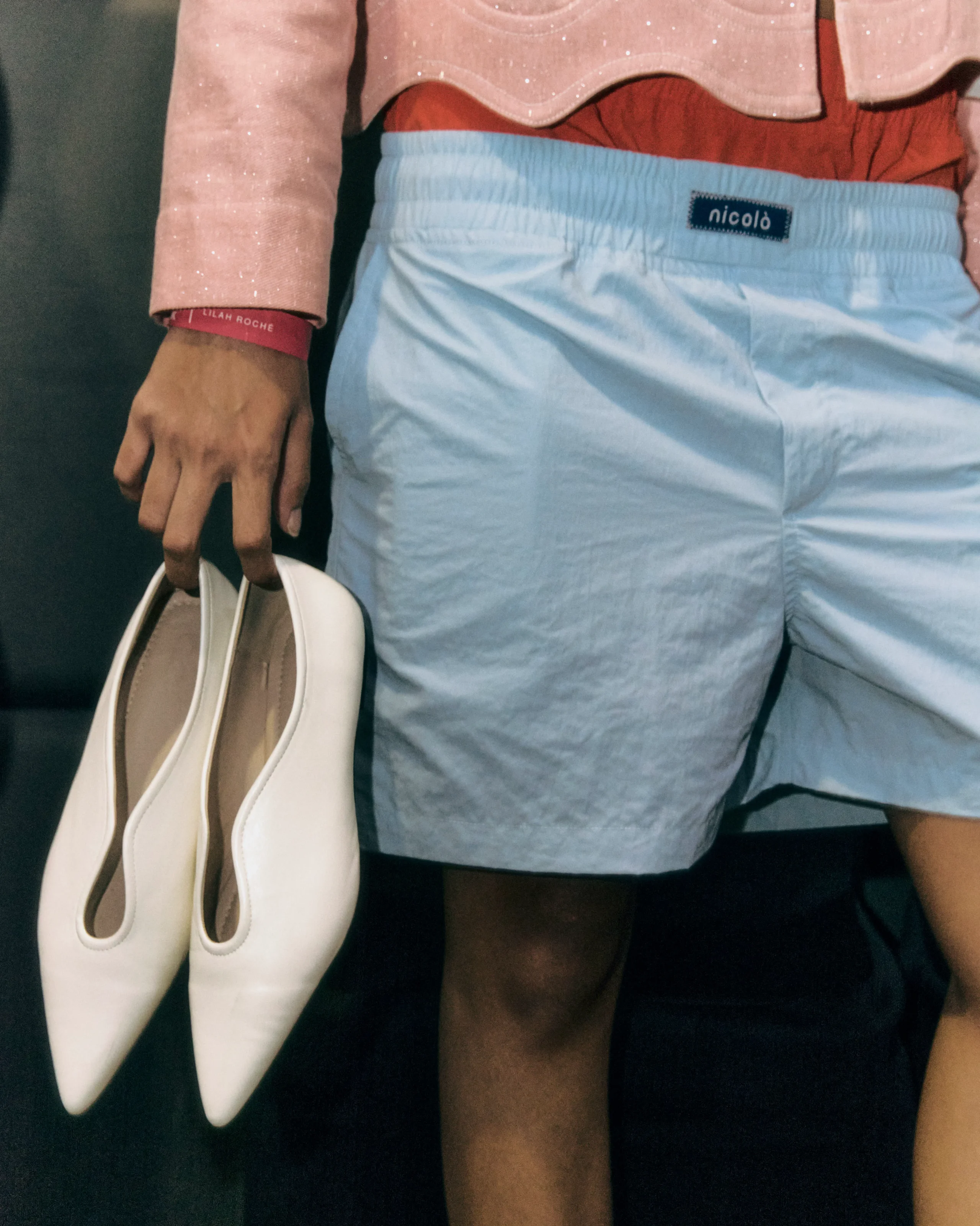 Person wearing light blue shorts and a pink sparkly top, holding white shoes; a pink bracelet with small white text is visible on the wrist.