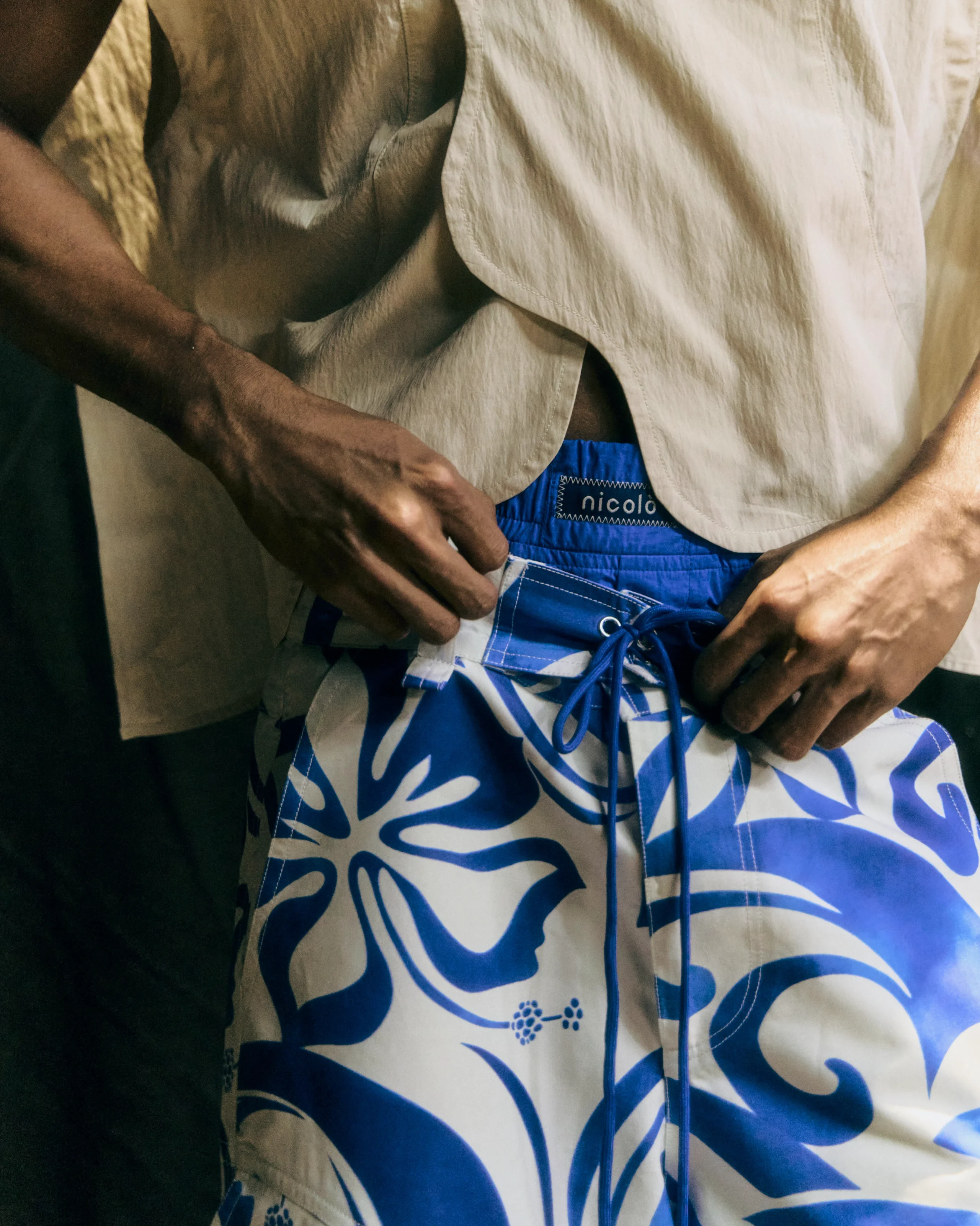 Close-up of a person adjusting blue patterned shorts beneath a beige shirt, hands visible.