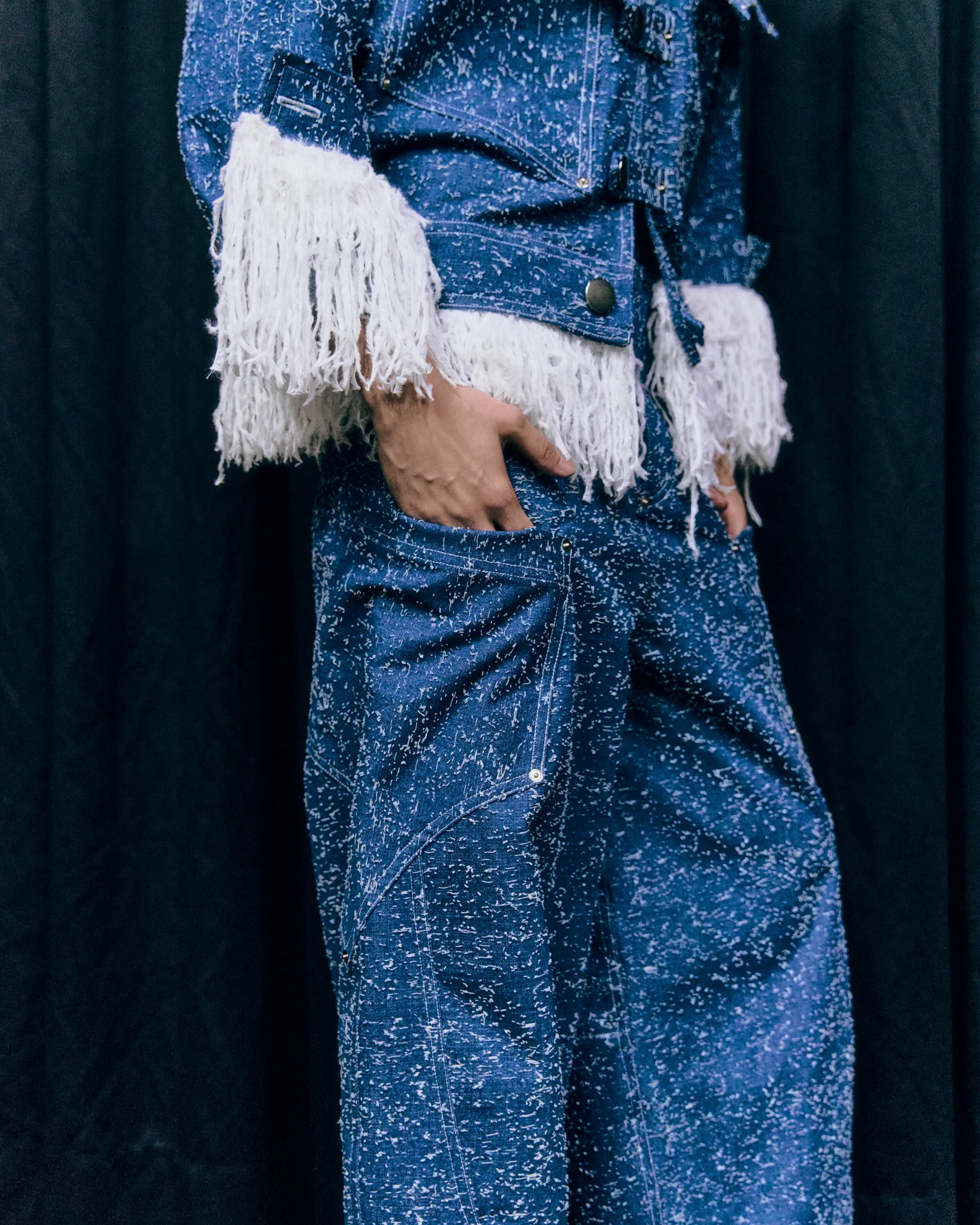 Close-up of a blue denim jacket with long white fringe sleeves against a dark background, showing the texture and splatter pattern of the fabric.