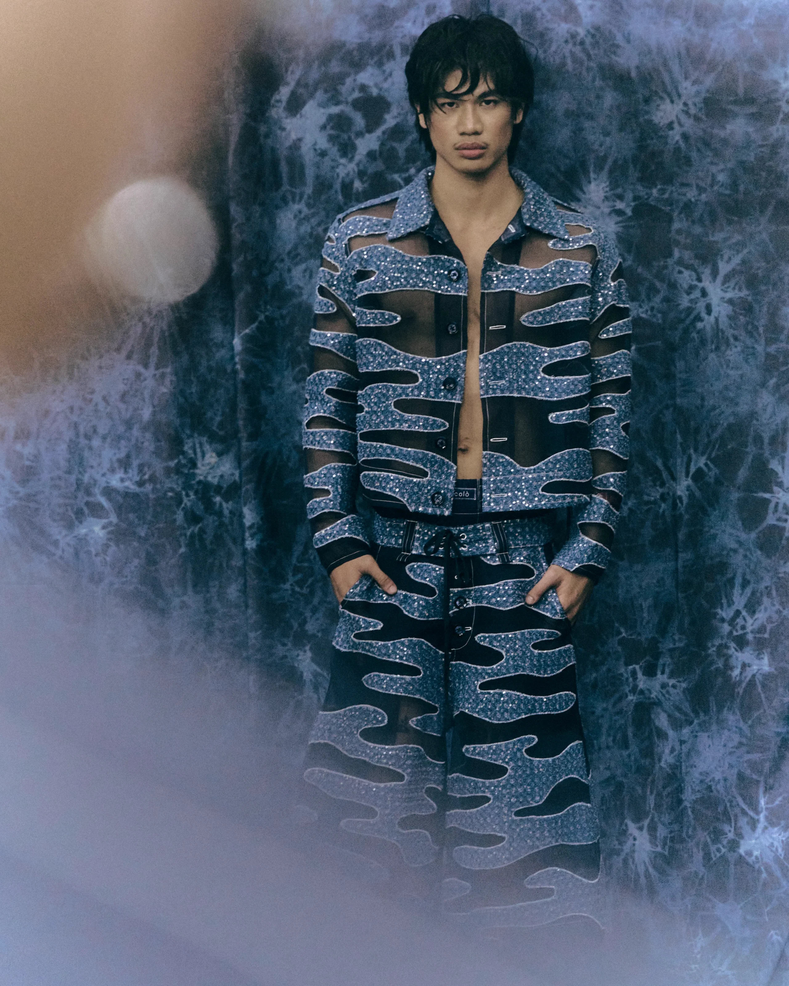 Fashion portrait of a young man in a sparkling blue cut-out jacket standing against a blue marbled backdrop.