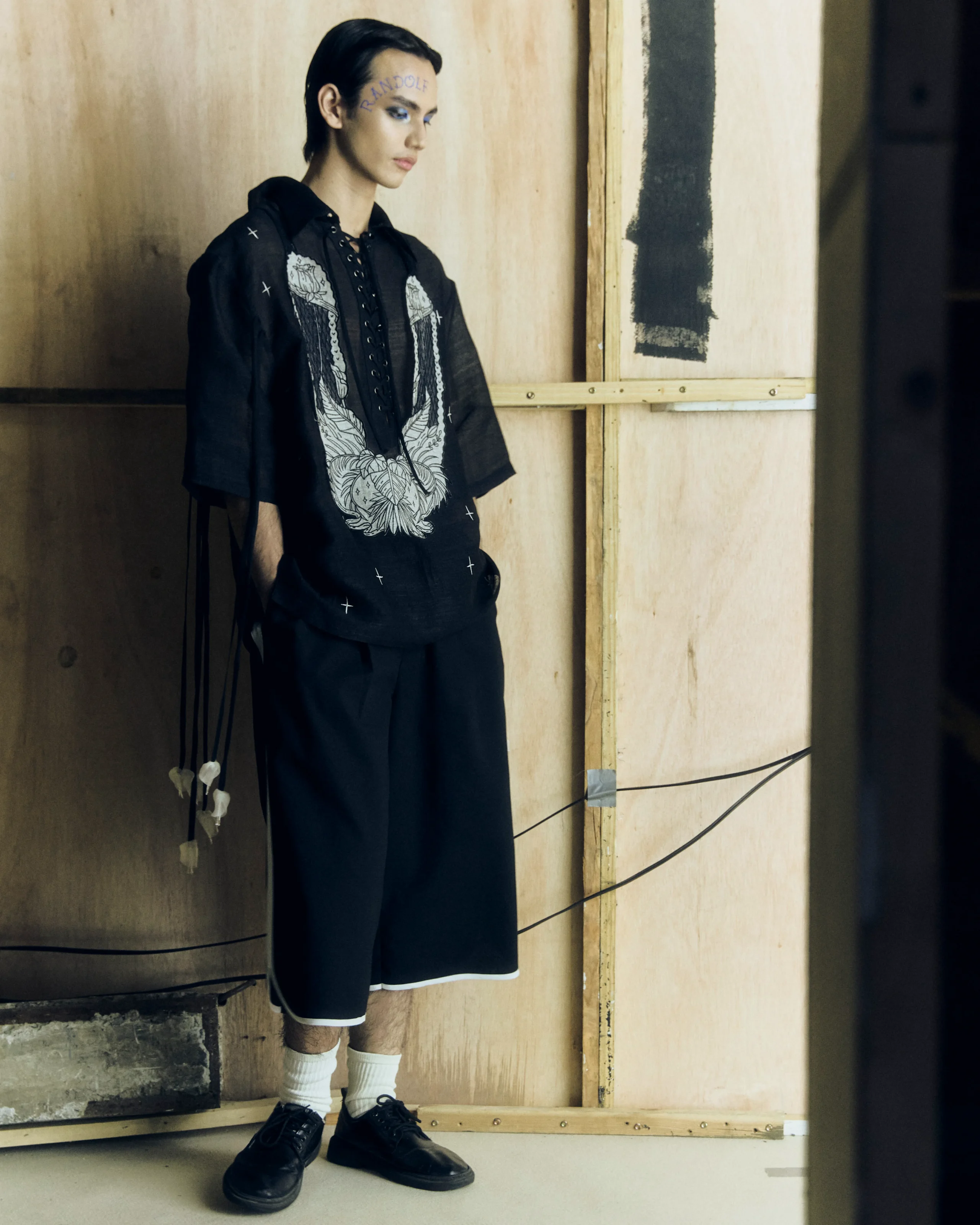 Person with short dark hair in a black embroidered outfit stands in a wooden studio setting, facing left in a side profile.