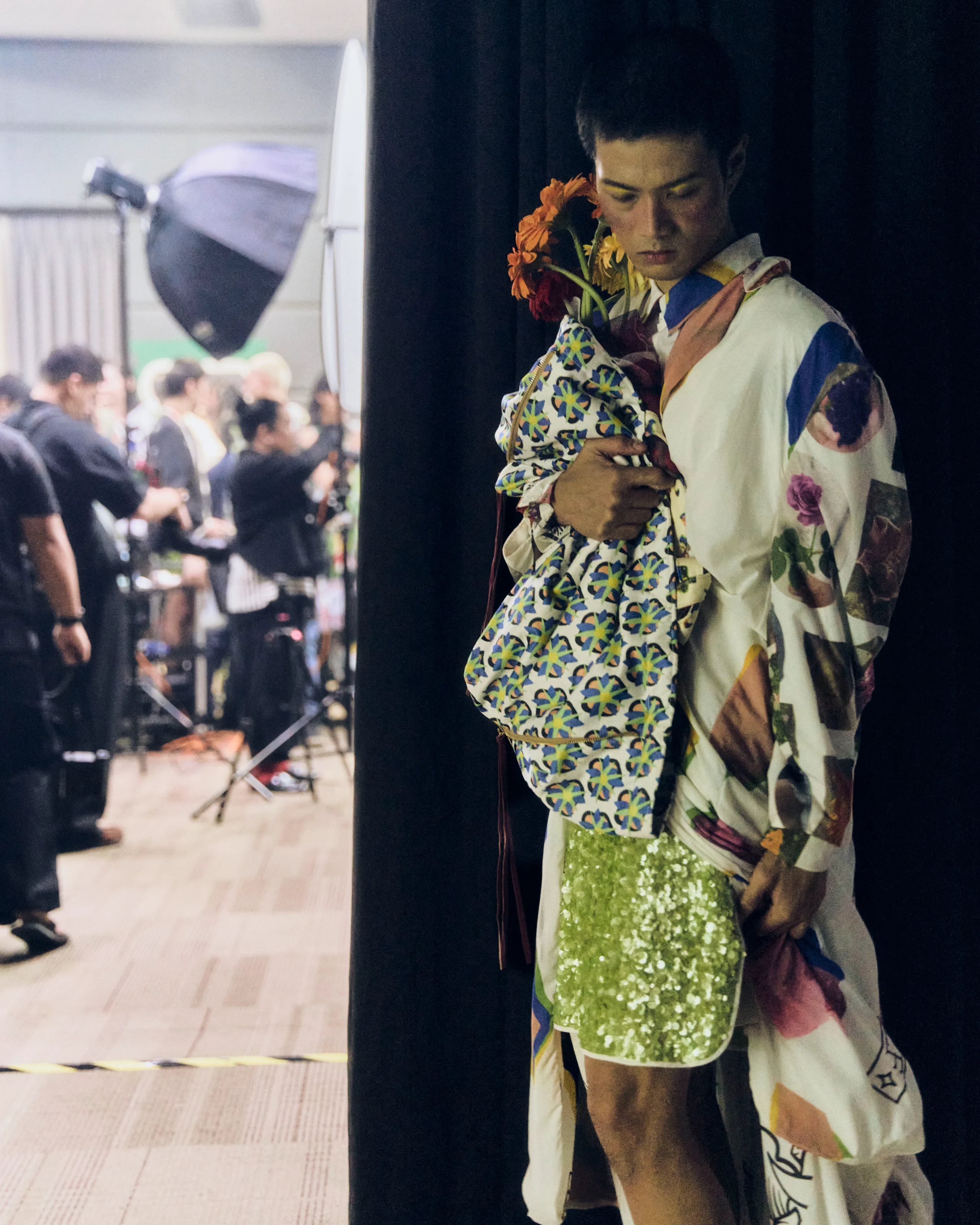Person backstage in a colorful patterned kimono, clutching a bouquet of orange flowers near a dark curtain.