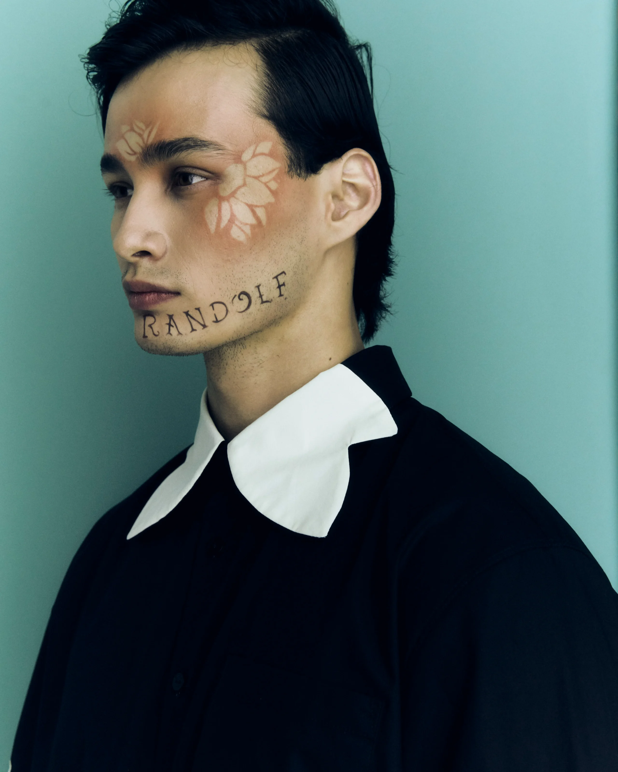 Young man in a black suit with white collar, side profile, teal background; pale floral face paint on the left cheek and 'RANDOLF' tattooed across the chin area.
