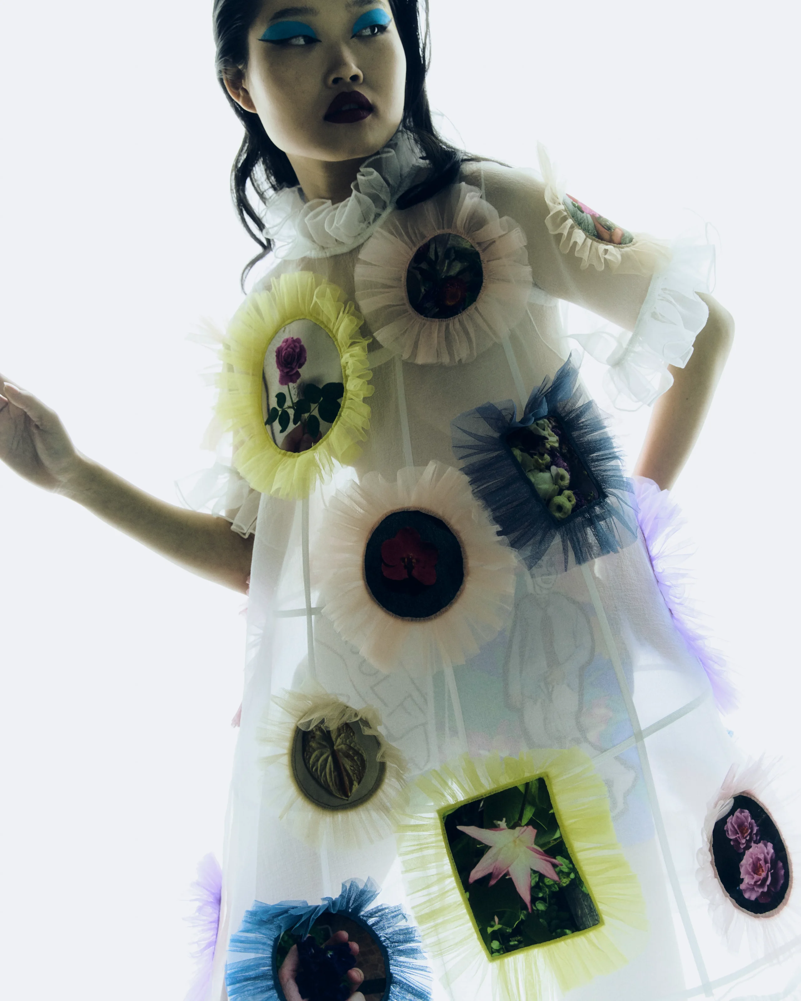 Model in a sheer white dress with large circular fabric patches and ruffled edges, floral embroidery visible at patches.