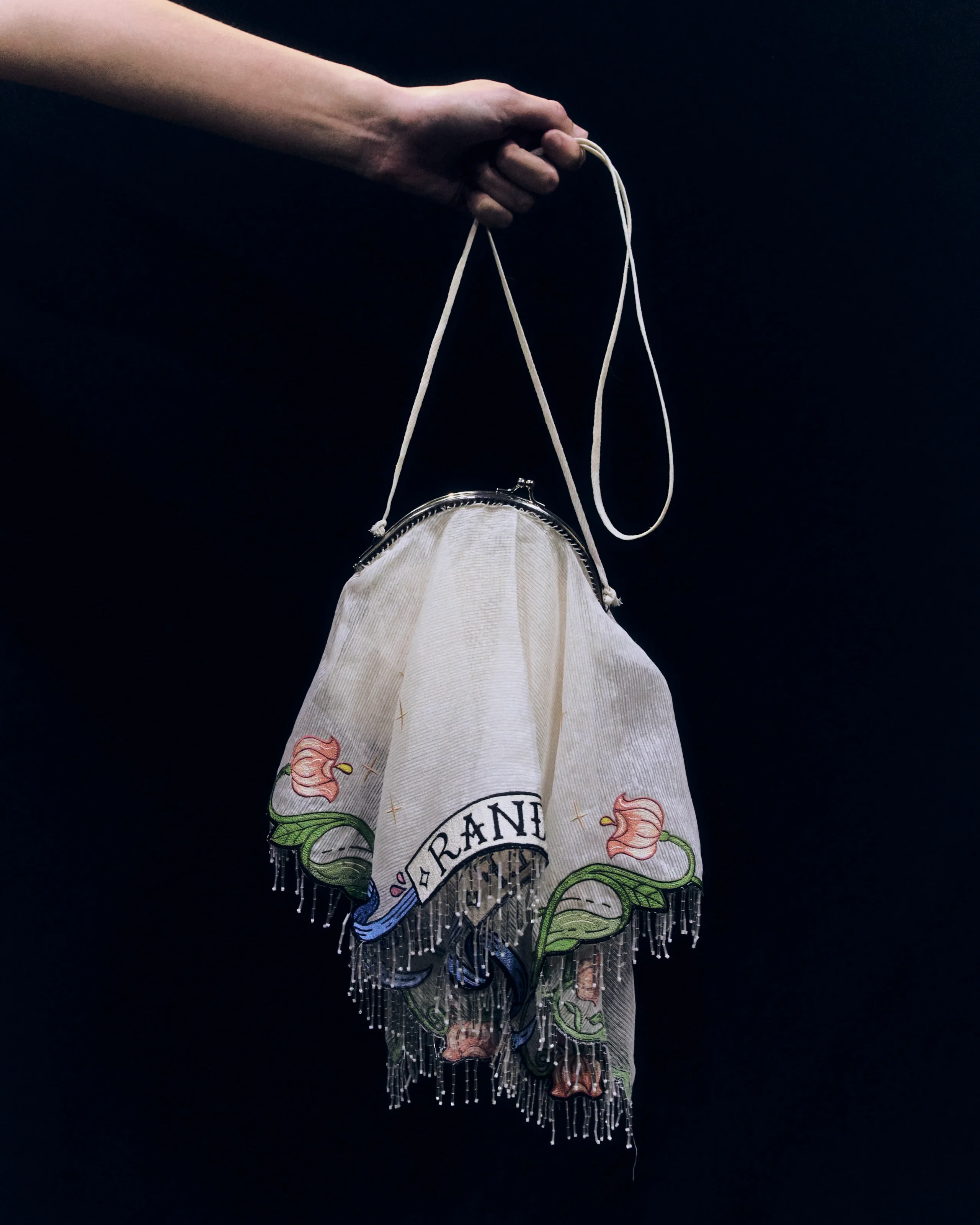 Close-up of a pale arm gripping a beige fringed scarf-like bag against a dark background, showcasing embroidered pink flowers and a circular emblem near the bottom edge.