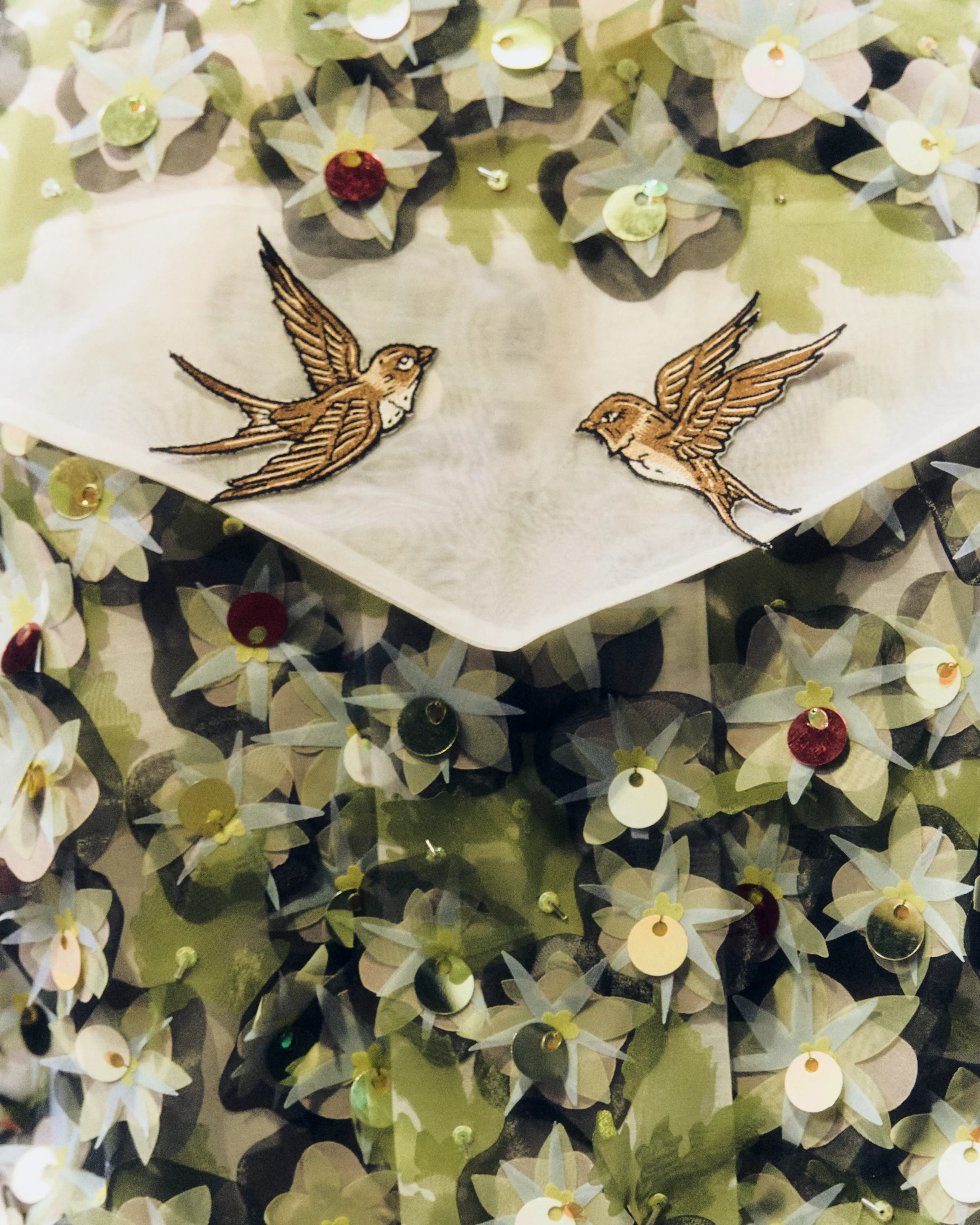 Two embroidered brown birds in flight on pale fabric, surrounded by green leaves and sequined circles.