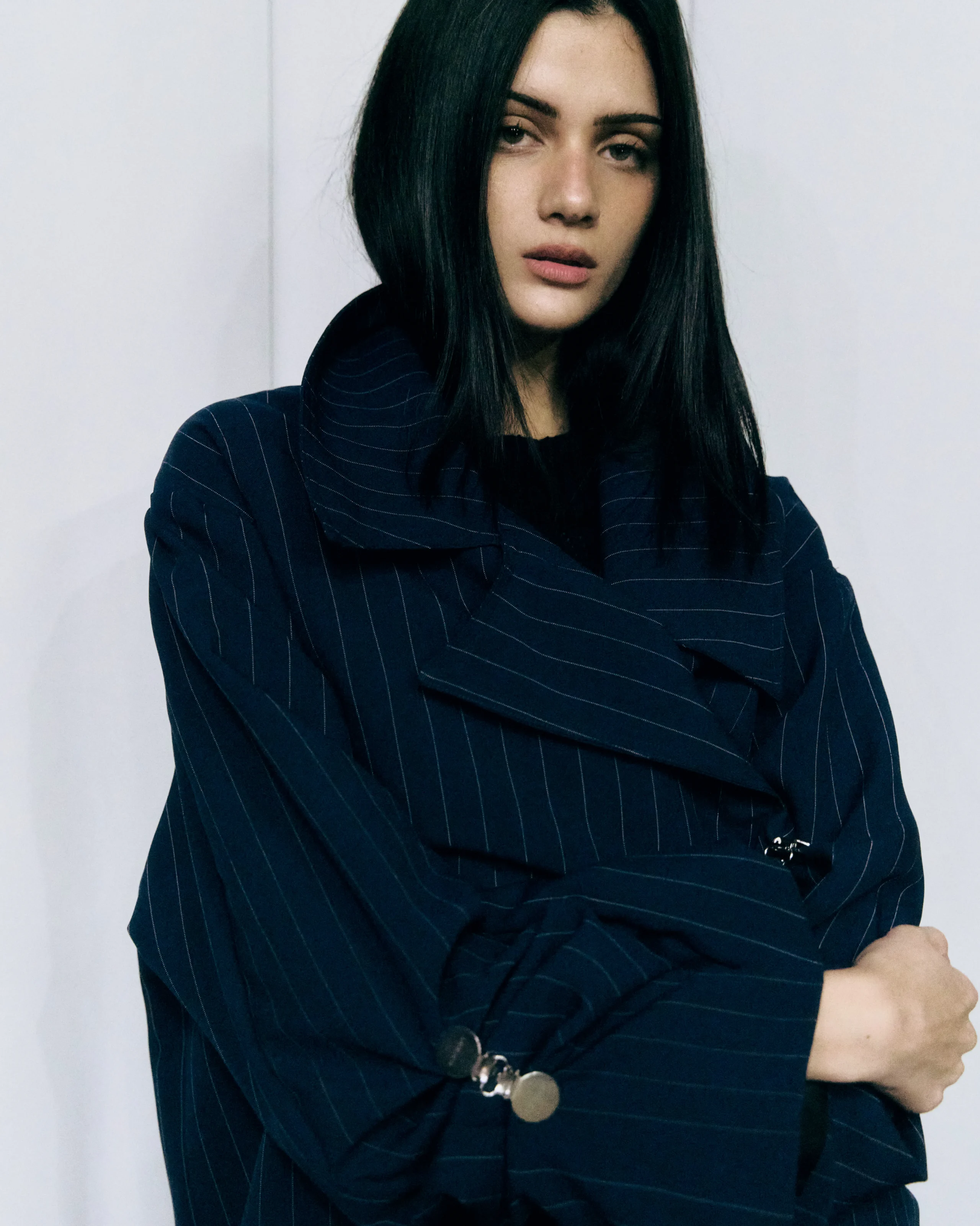Woman with long dark hair wearing a navy pinstripe blazer, arms crossed, looking at the camera against a light backdrop.