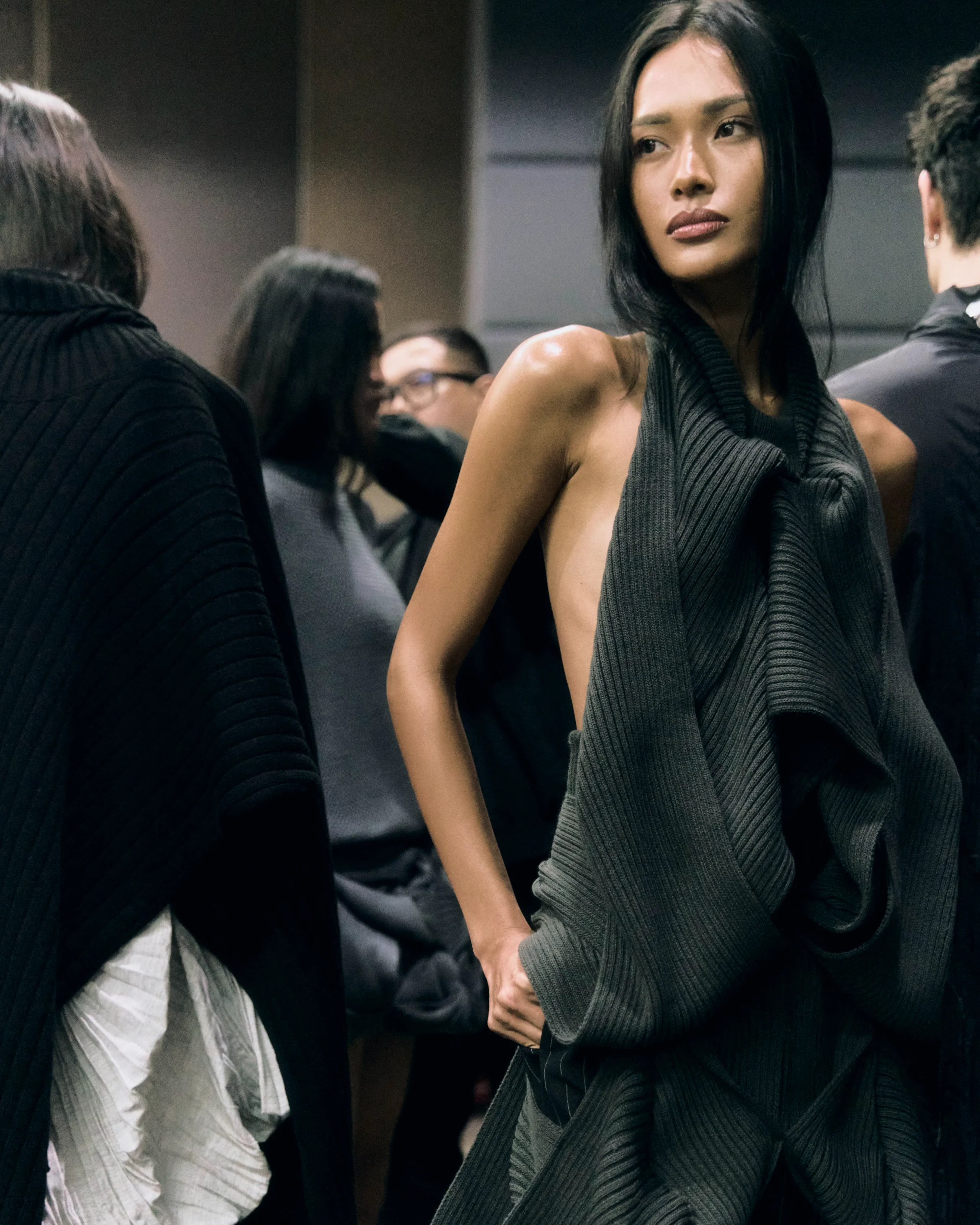 Model in a dark grey ribbed dress, looking to the side, surrounded by people in a backstage setting.