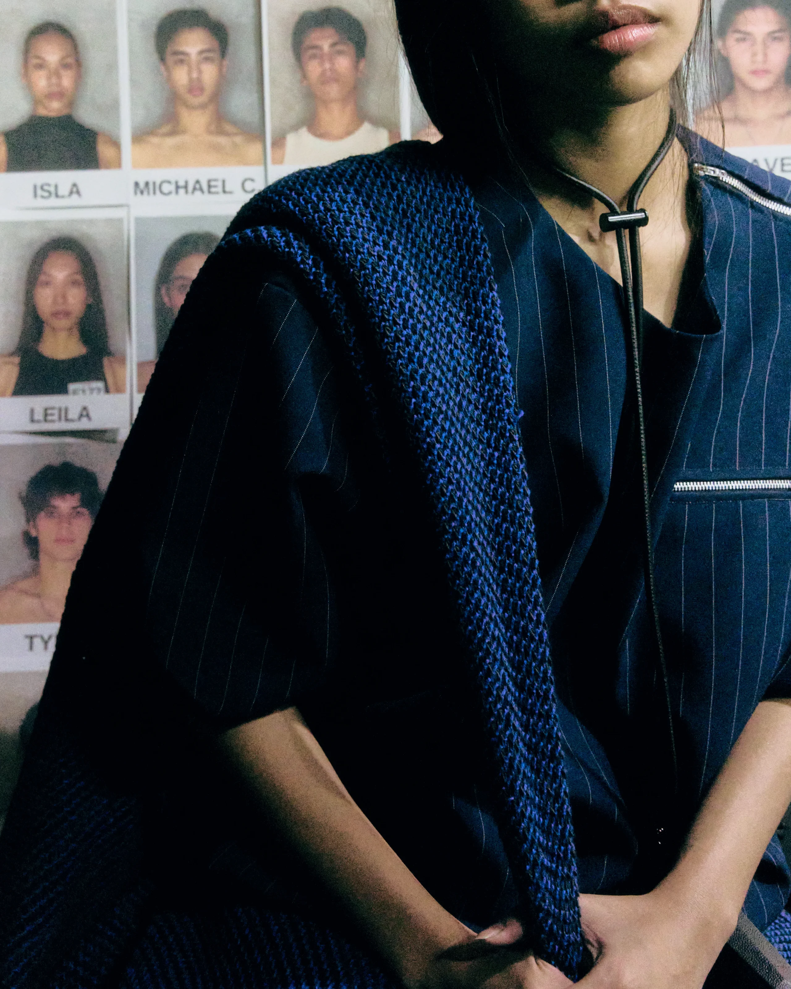 Person wearing a blue pinstripe blazer and patterned scarf, standing in front of mugshot posters on a wall, cropped to chest level.