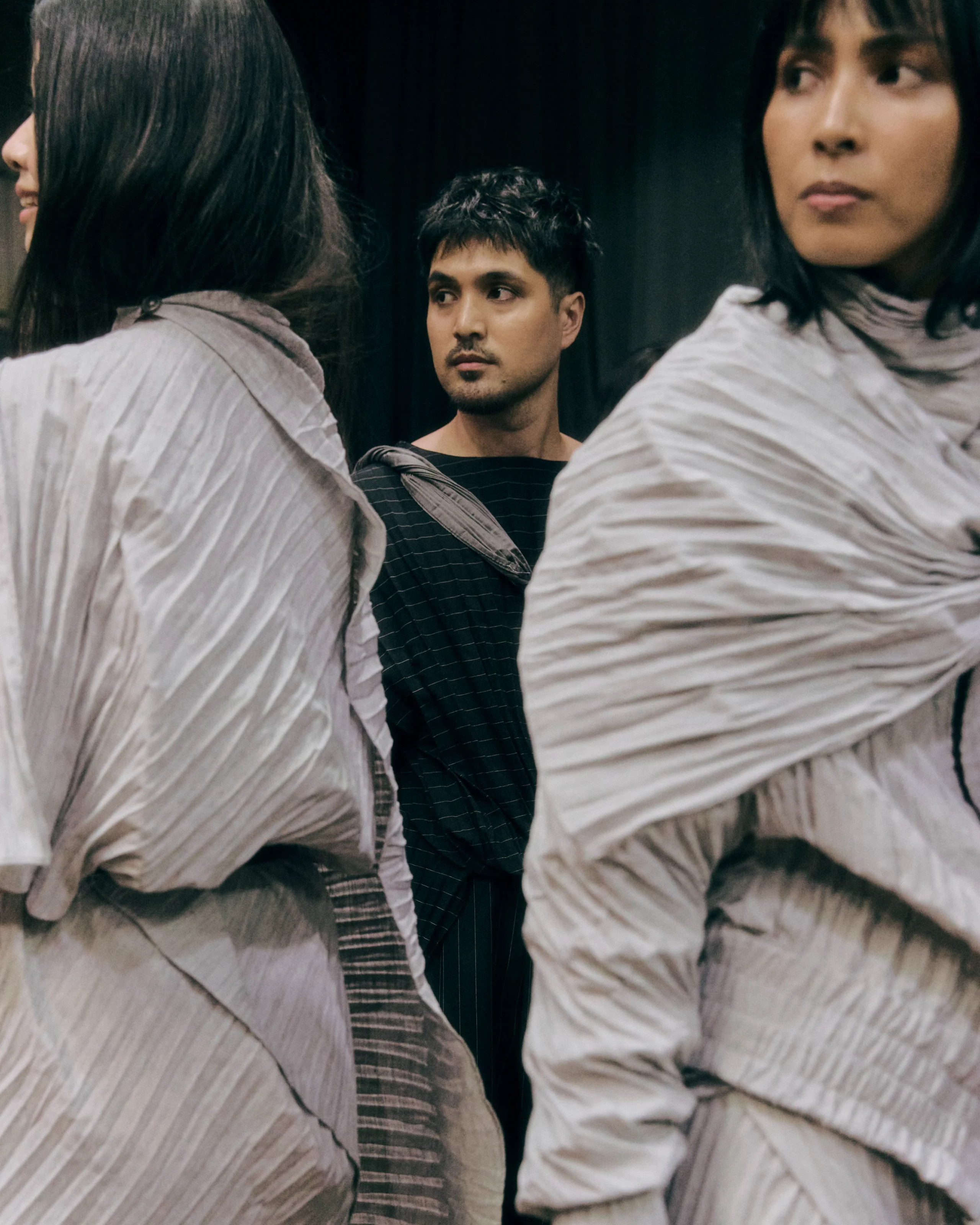 Three people in neutral-toned clothing, backs turned; a man in the center looks to his left with a contemplative expression.