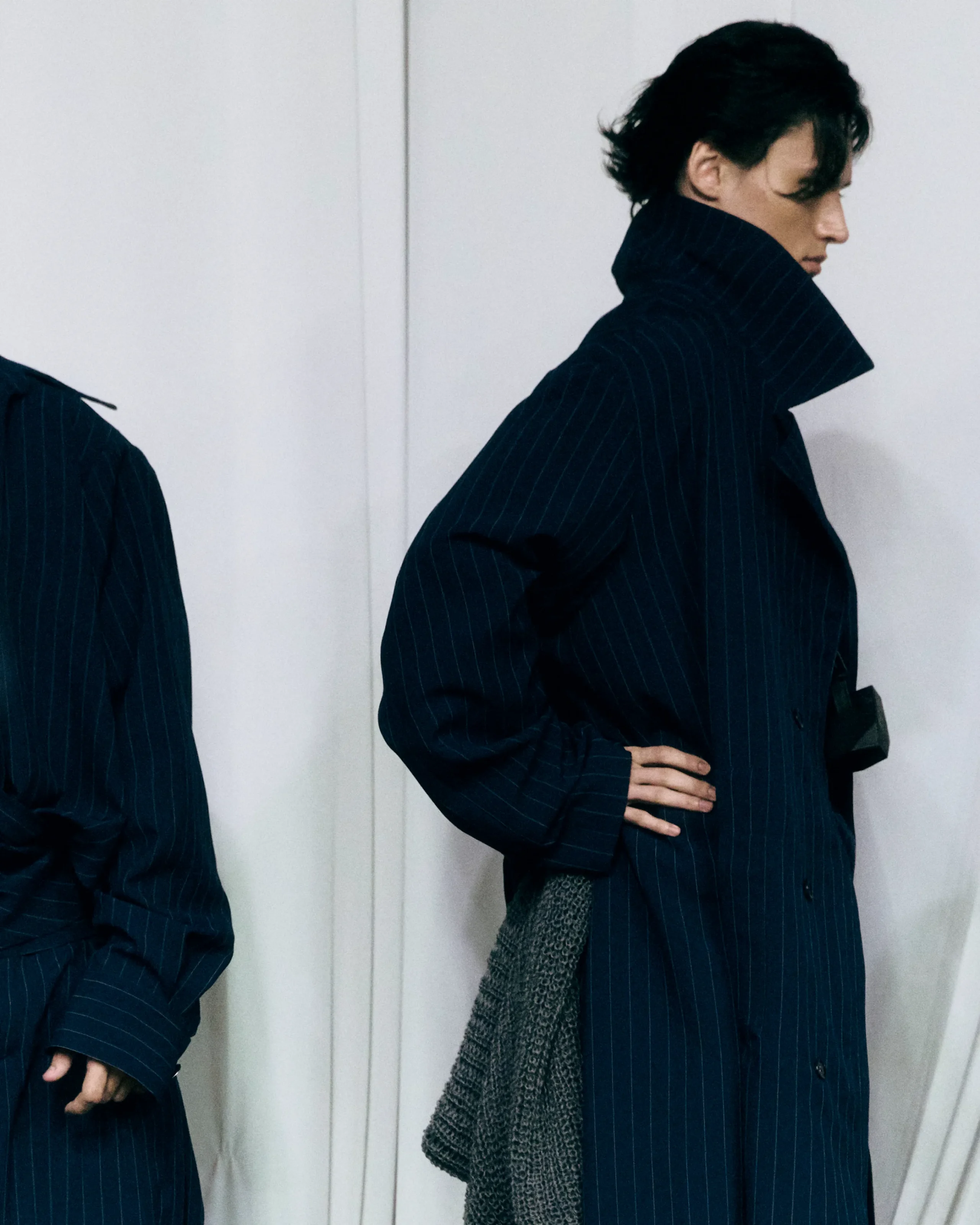 Side profile of a person wearing a dark pinstripe coat with a high collar, hand on hip, holding a gray knit bag.