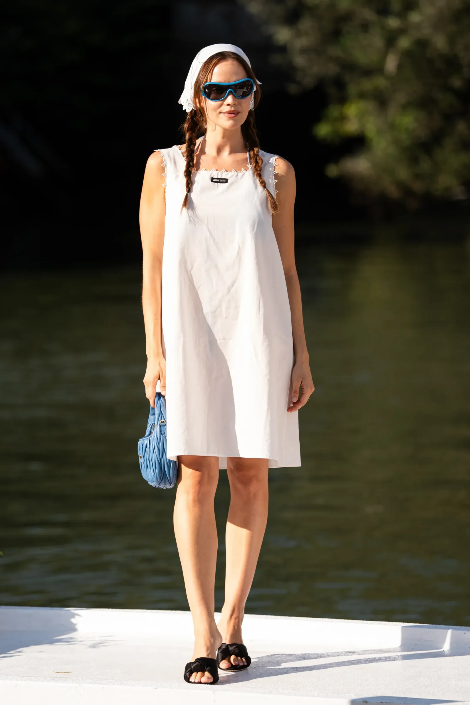 Woman standing by water in a white sleeveless dress, wearing a white headscarf and blue sunglasses, with braided hair and a blue bag hanging at her side.