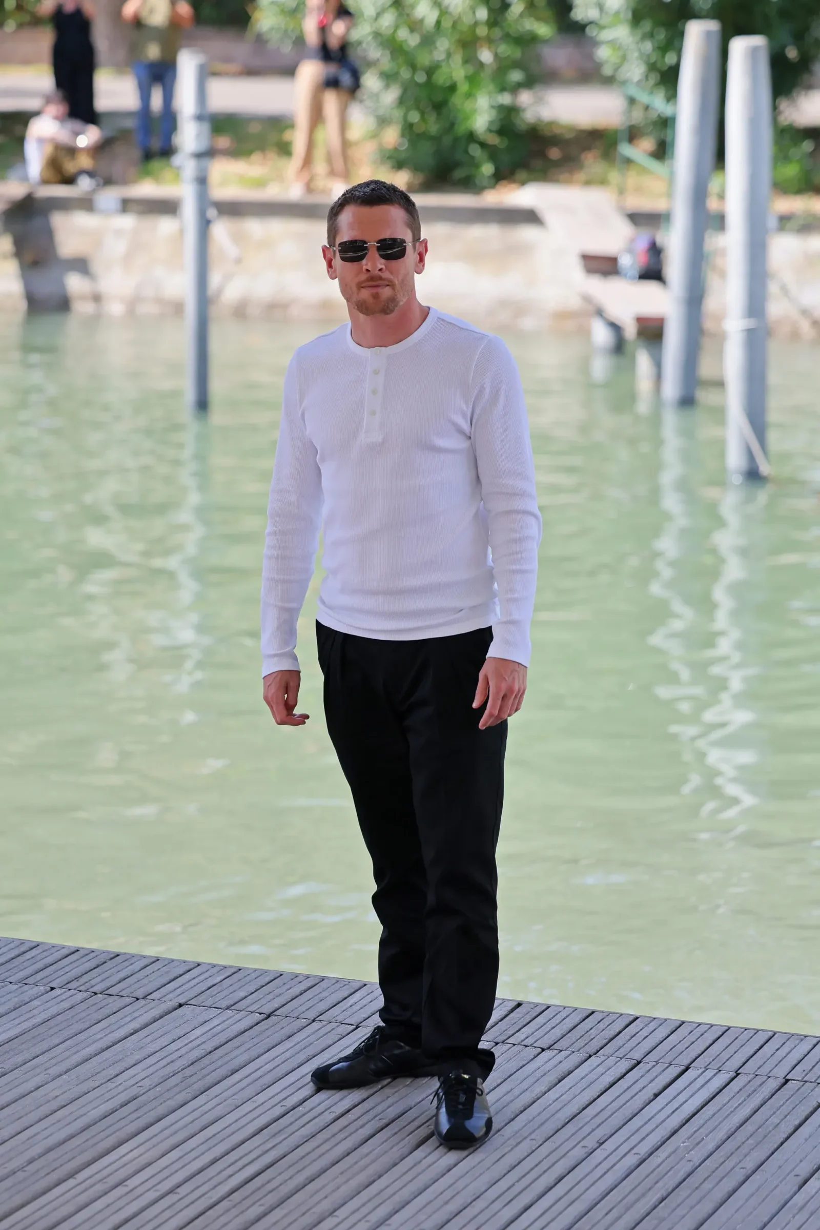 Man in a white long-sleeve shirt and black pants stands on a wooden dock beside a greenish body of water, wearing sunglasses.