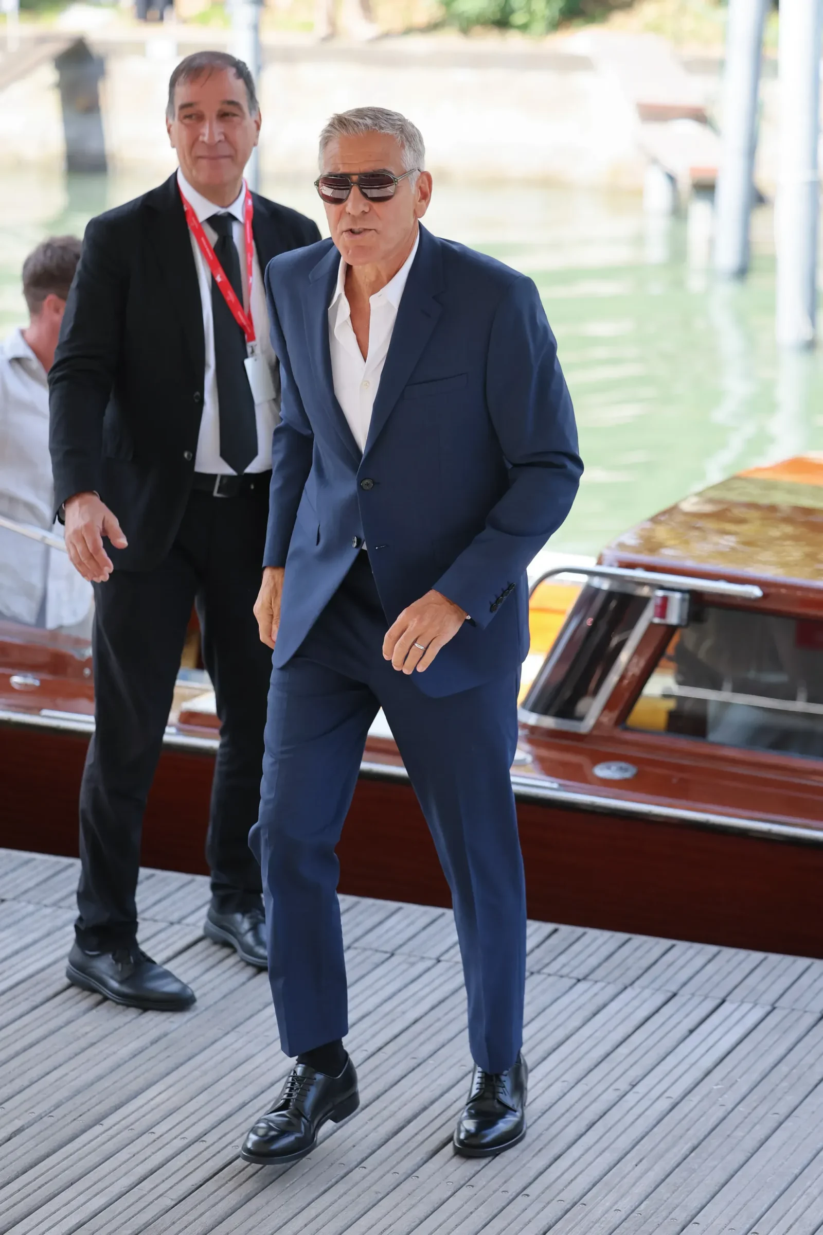 Man in a blue suit and sunglasses steps onto a docked boat, with another suited man in the background wearing a red lanyard.