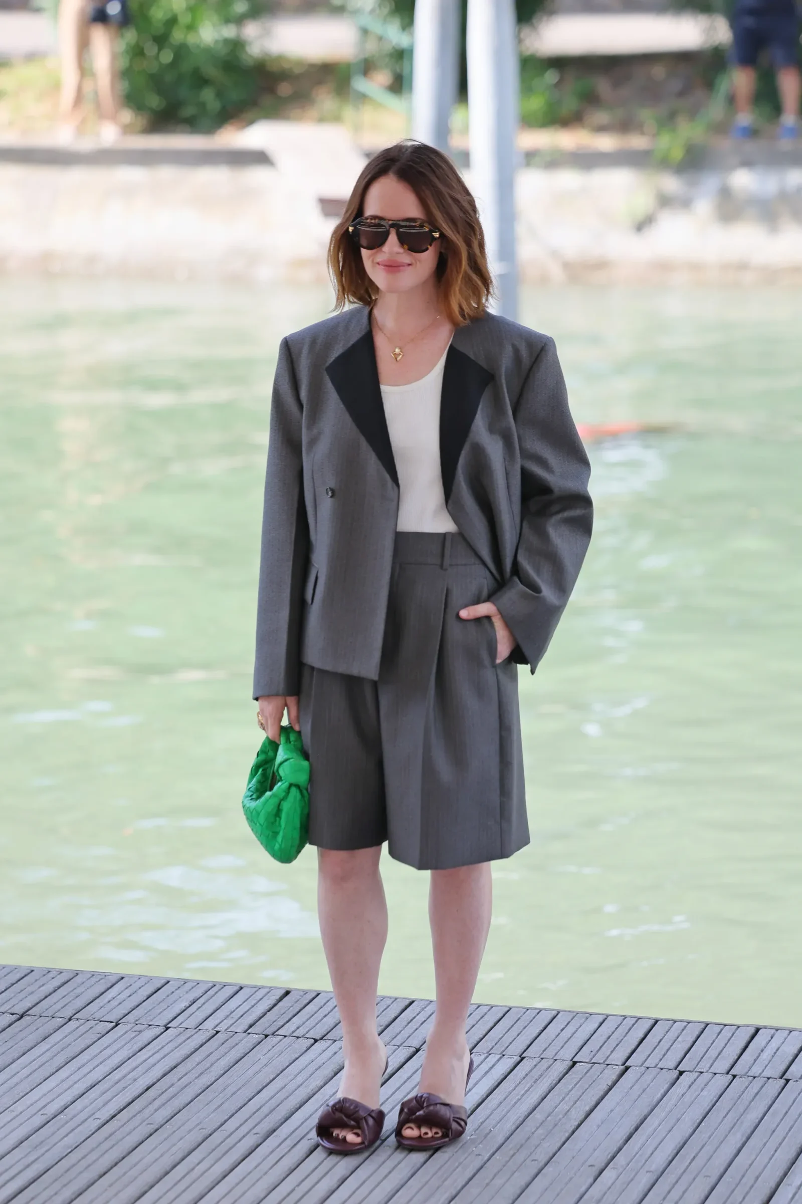 Woman in gray blazer and matching shorts, white top, sunglasses, standing on a wooden dock by greenish water, holding a bright green handbag.