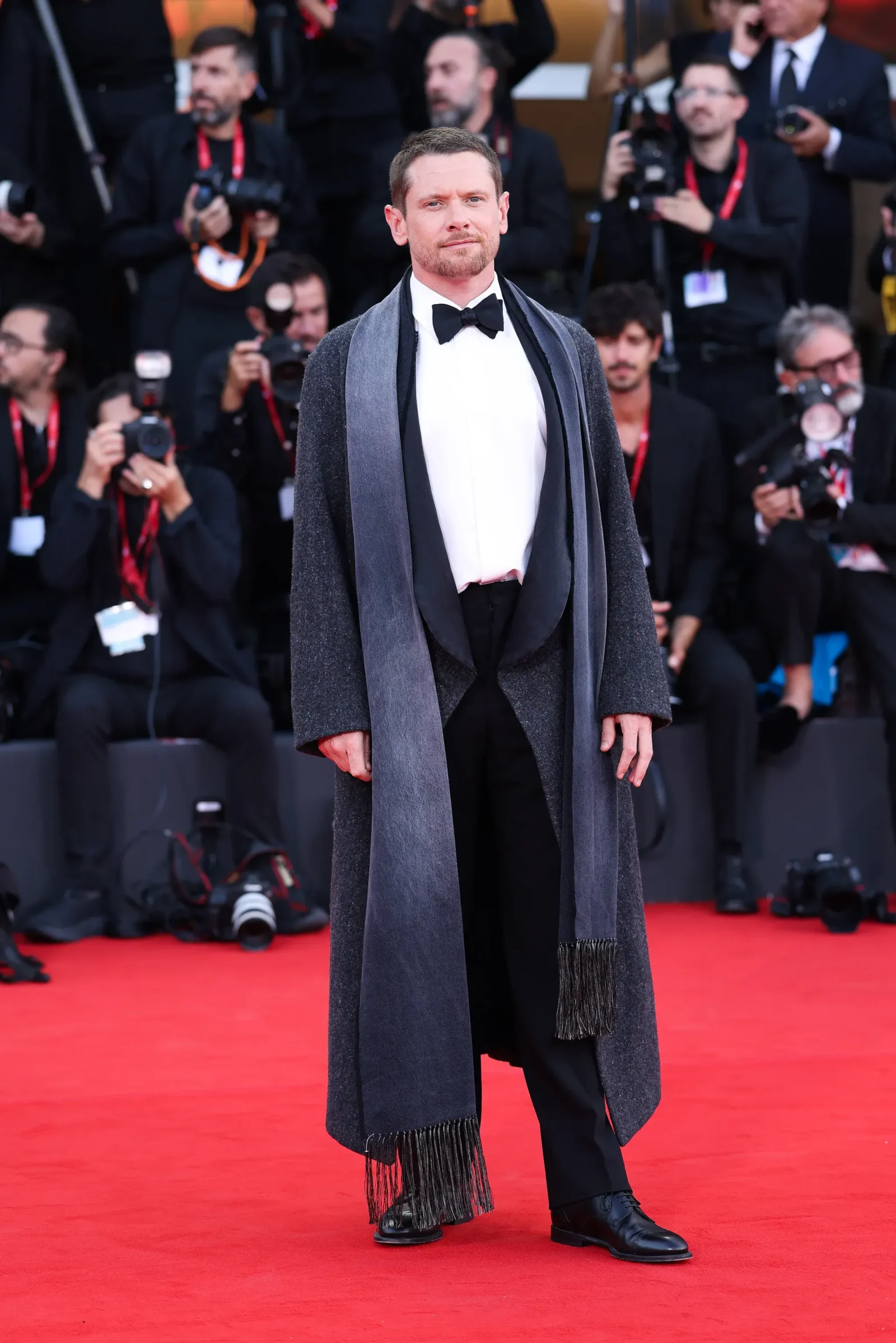 Man in tuxedo and gray scarf poses on red carpet as photographers look on, behind him. Guards and camera crews in the background.