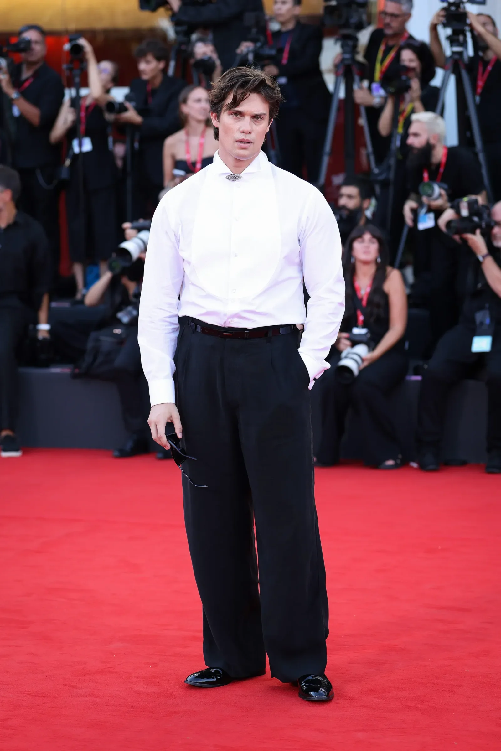 Man in a white dress shirt and black trousers stands on a red carpet as paparazzi photograph him from behind, holding sunglasses in one hand.