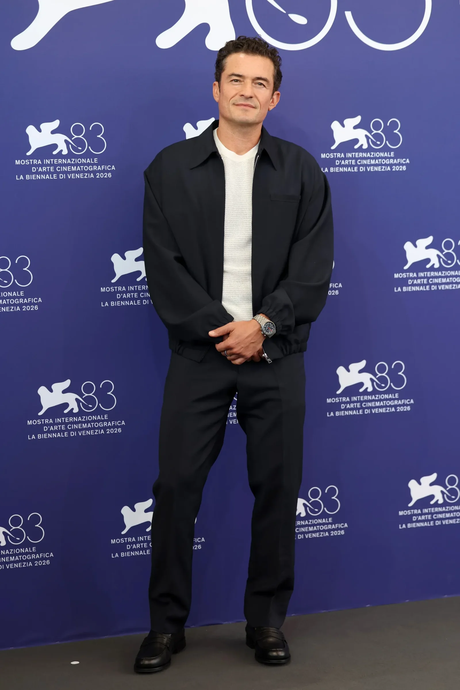 Man in a black jacket and white knit shirt stands with hands clasped in front, posed before a blue Venice Biennale backdrop with logos.