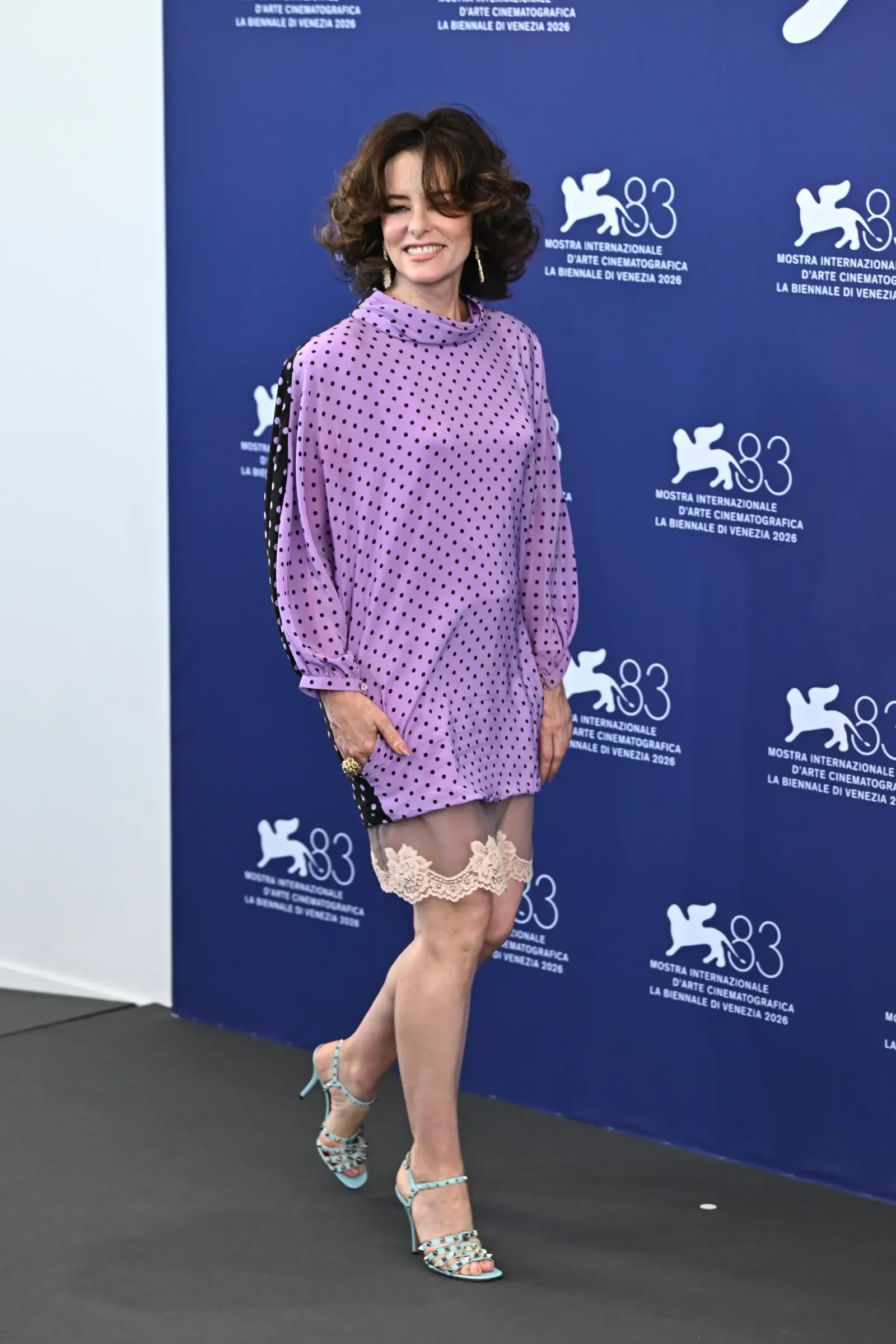 Woman posing on a blue backdrop with Biennale logos, wearing a lavender polka-dot dress, lace stockings, and turquoise strappy heels.