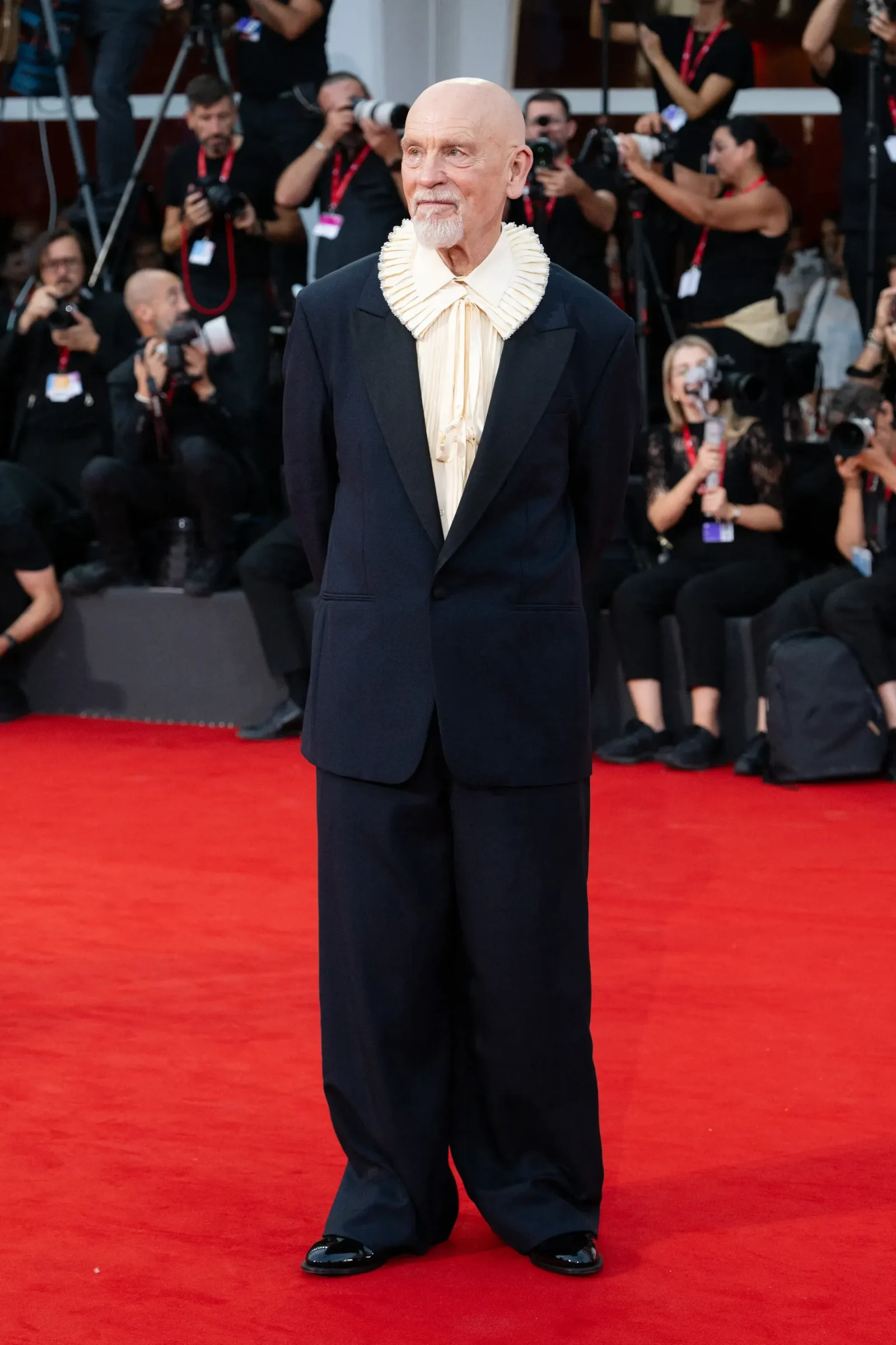 Man with a shaved head and goatee, wearing a dark suit with a cream ruffled shirt, standing on a red carpet as photographers photograph him in the background.