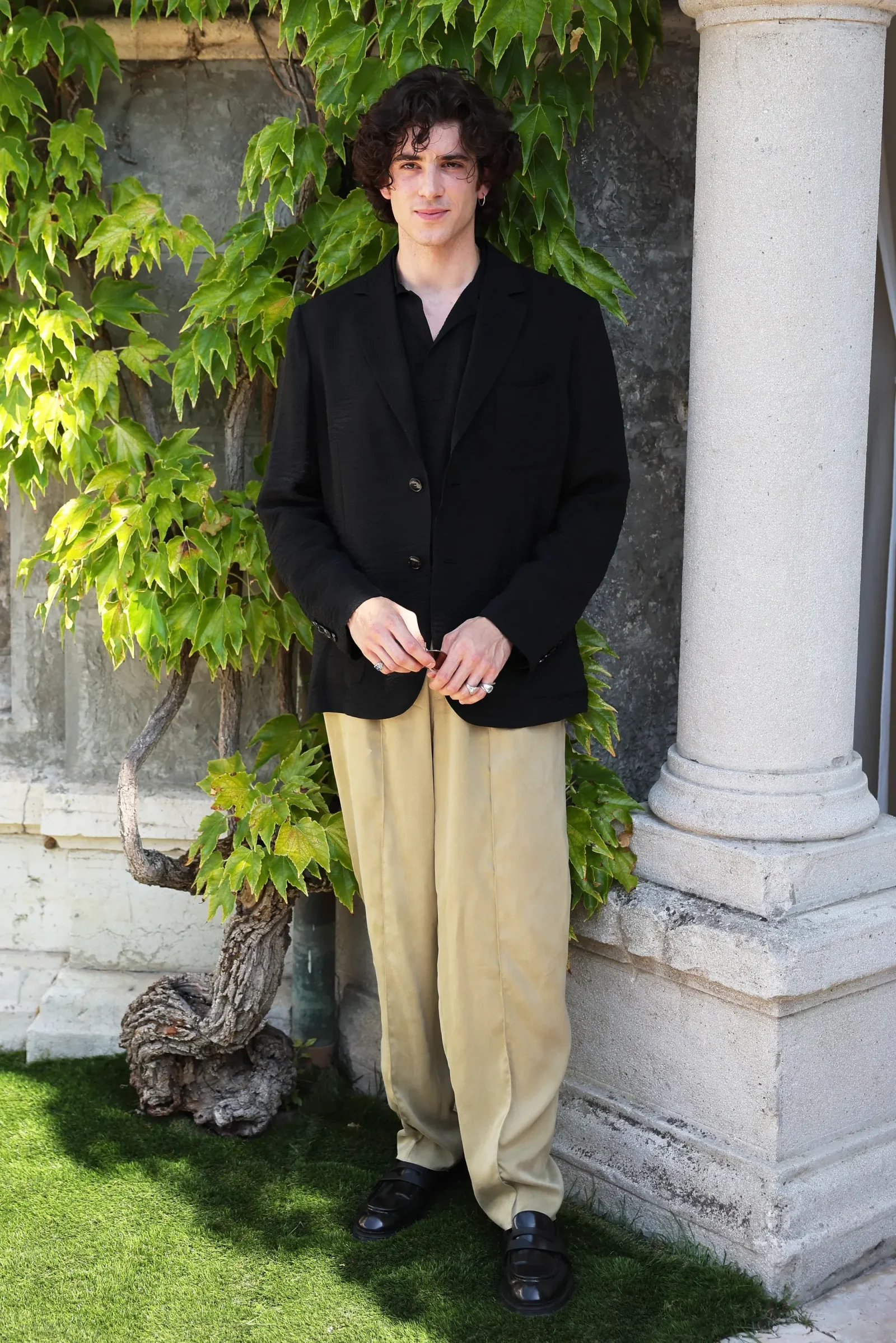 Young man standing outdoors beside a stone column, wearing a black blazer and beige wide-leg pants among ivy-covered walls.