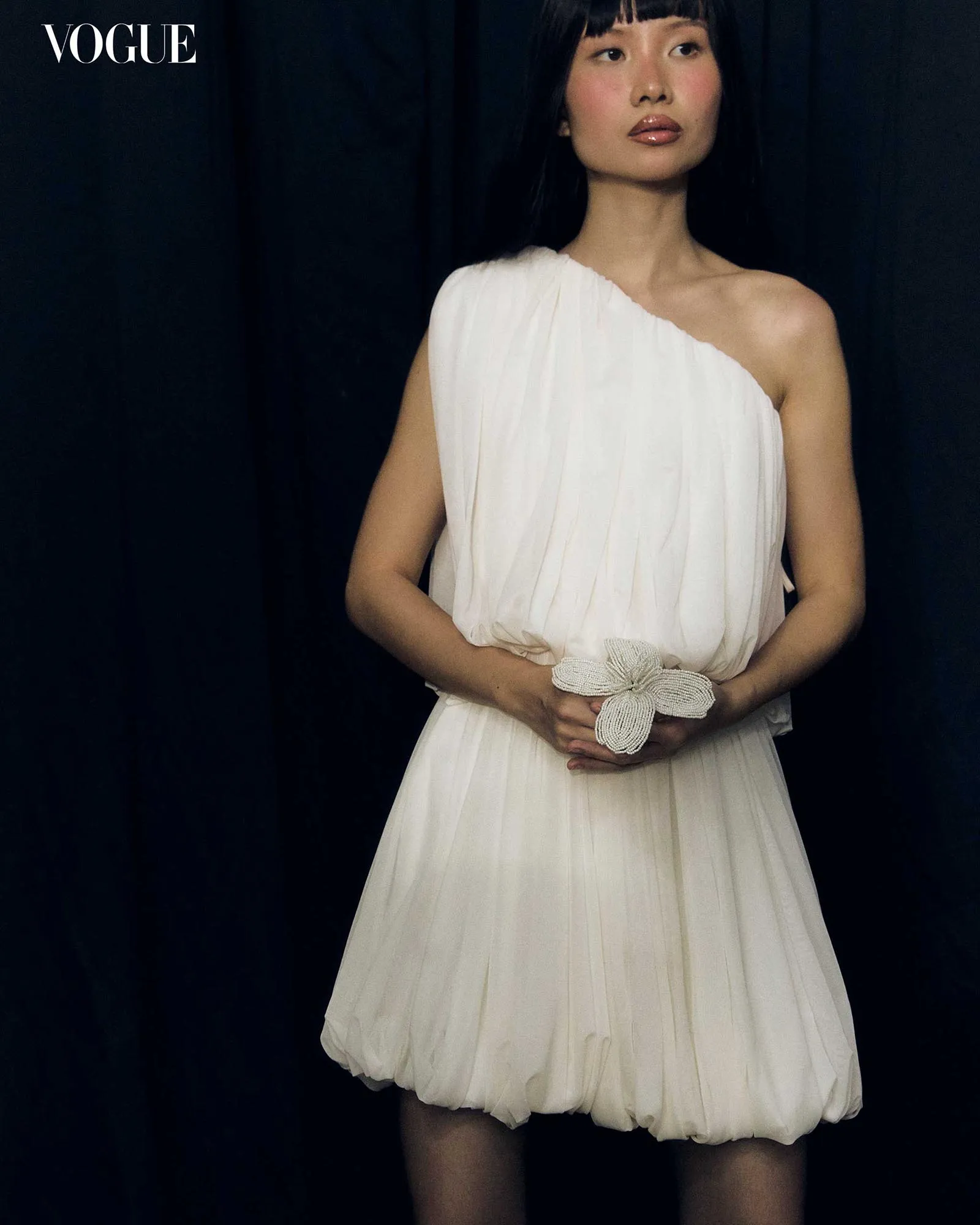Portrait of a woman in a white one-shoulder pleated dress, holding a beaded belt, against a dark backdrop with a Vogue logo in the top-left corner.