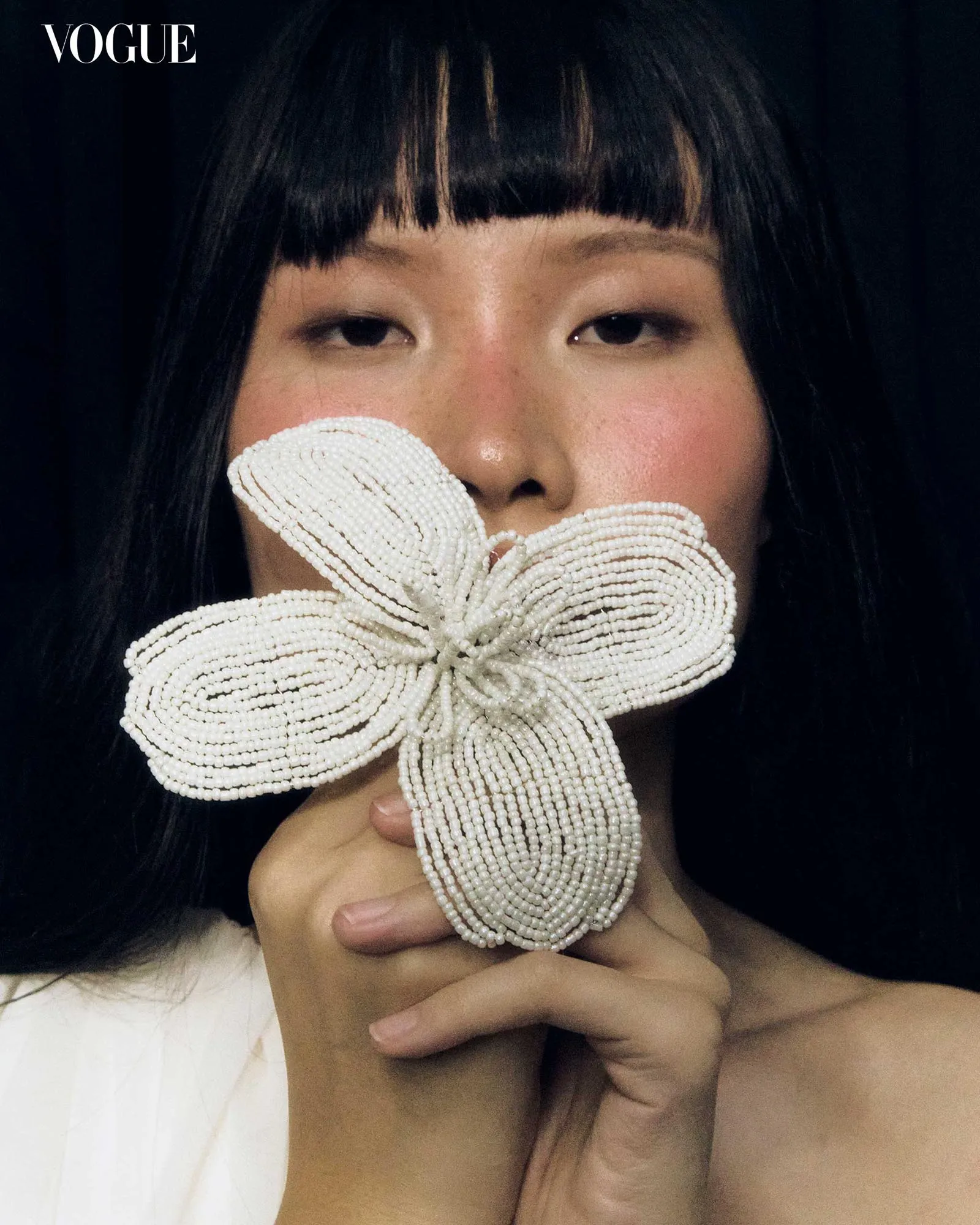 Close-up of a person with a large white beaded flower held over the mouth, Vogue logo visible in the top-left corner.