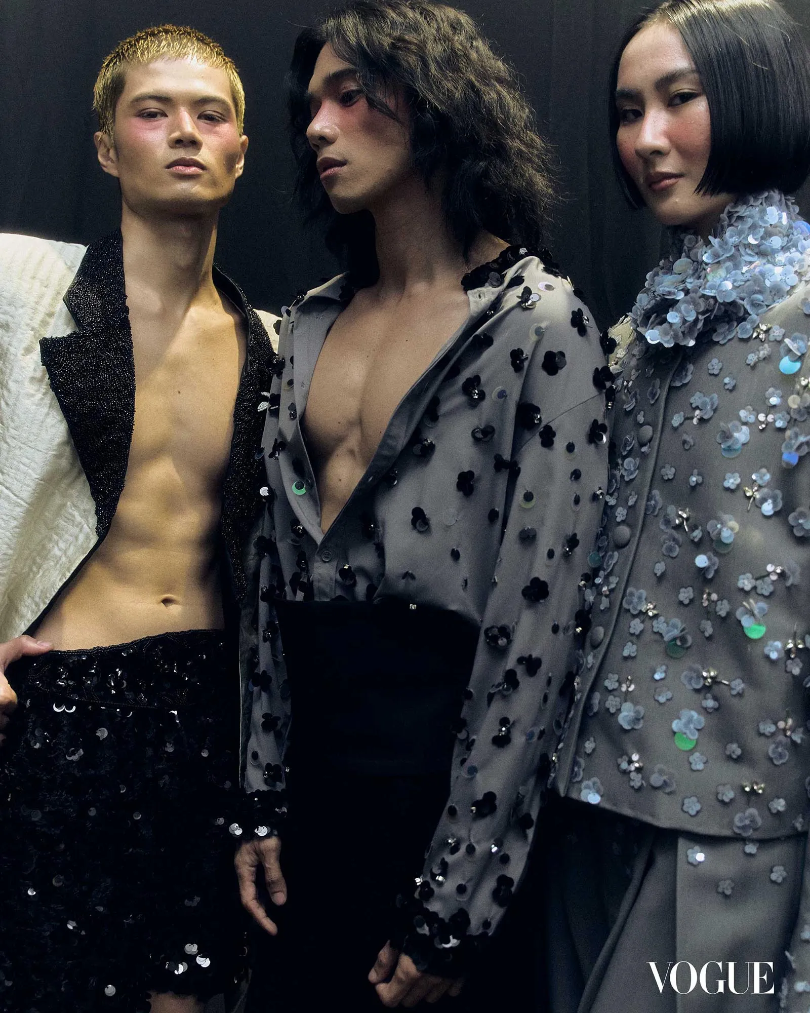 Three fashion models pose closely in sequined outfits with a dark backdrop; Vogue watermark visible in the bottom right.