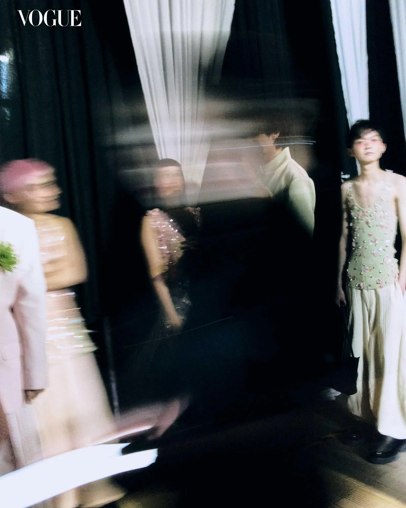 Vogue logo in the top-left corner of a backstage fashion show scene with blurred models in pastel gowns near dark curtains.
